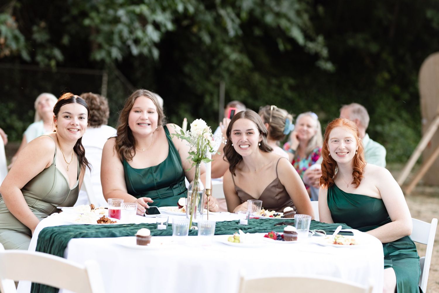 Four bridesmaids in green dresses smiling at a wedding reception table outdoors.