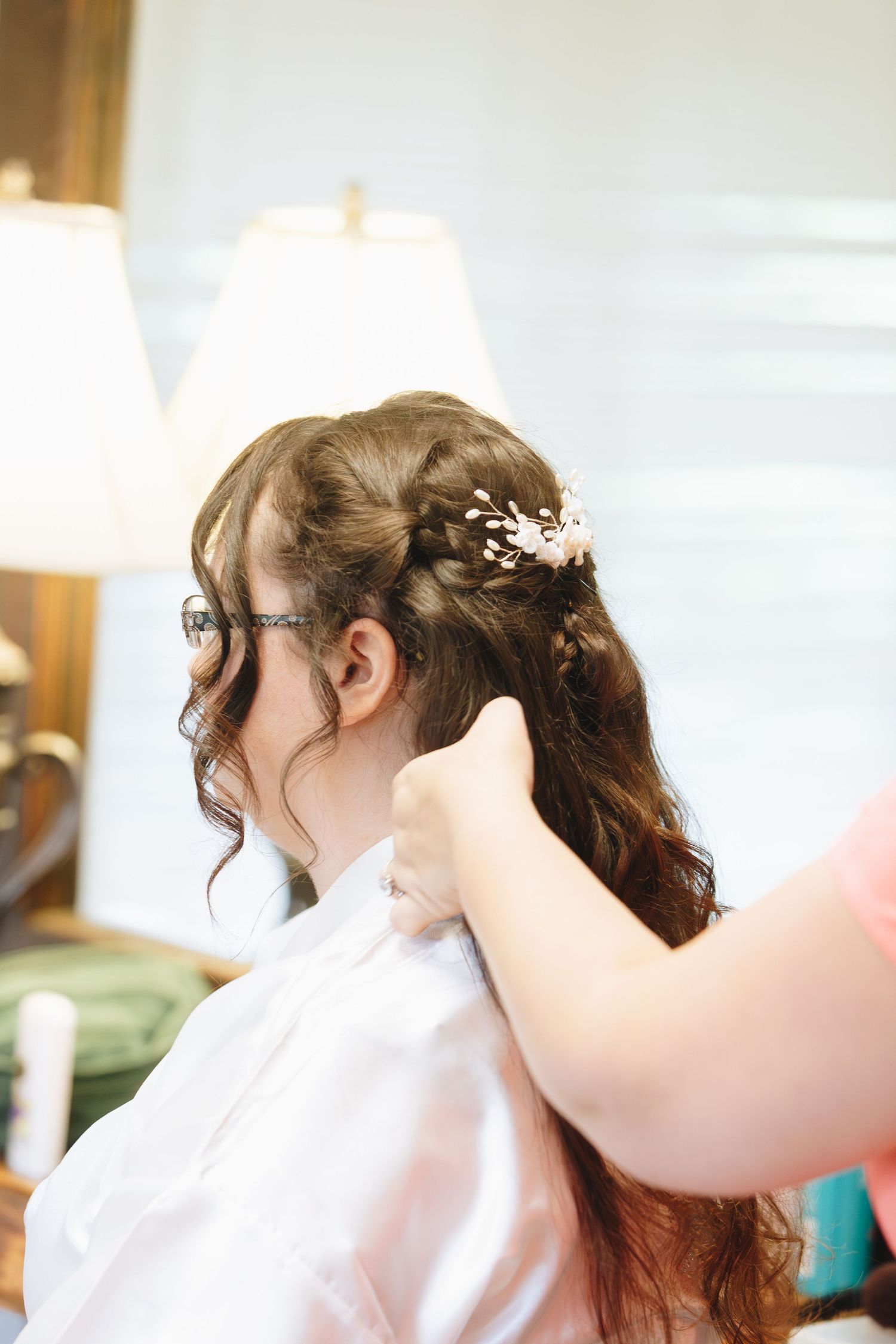 Hairstylist adding floral hair pin to bride's curly updo during wedding preparation.
