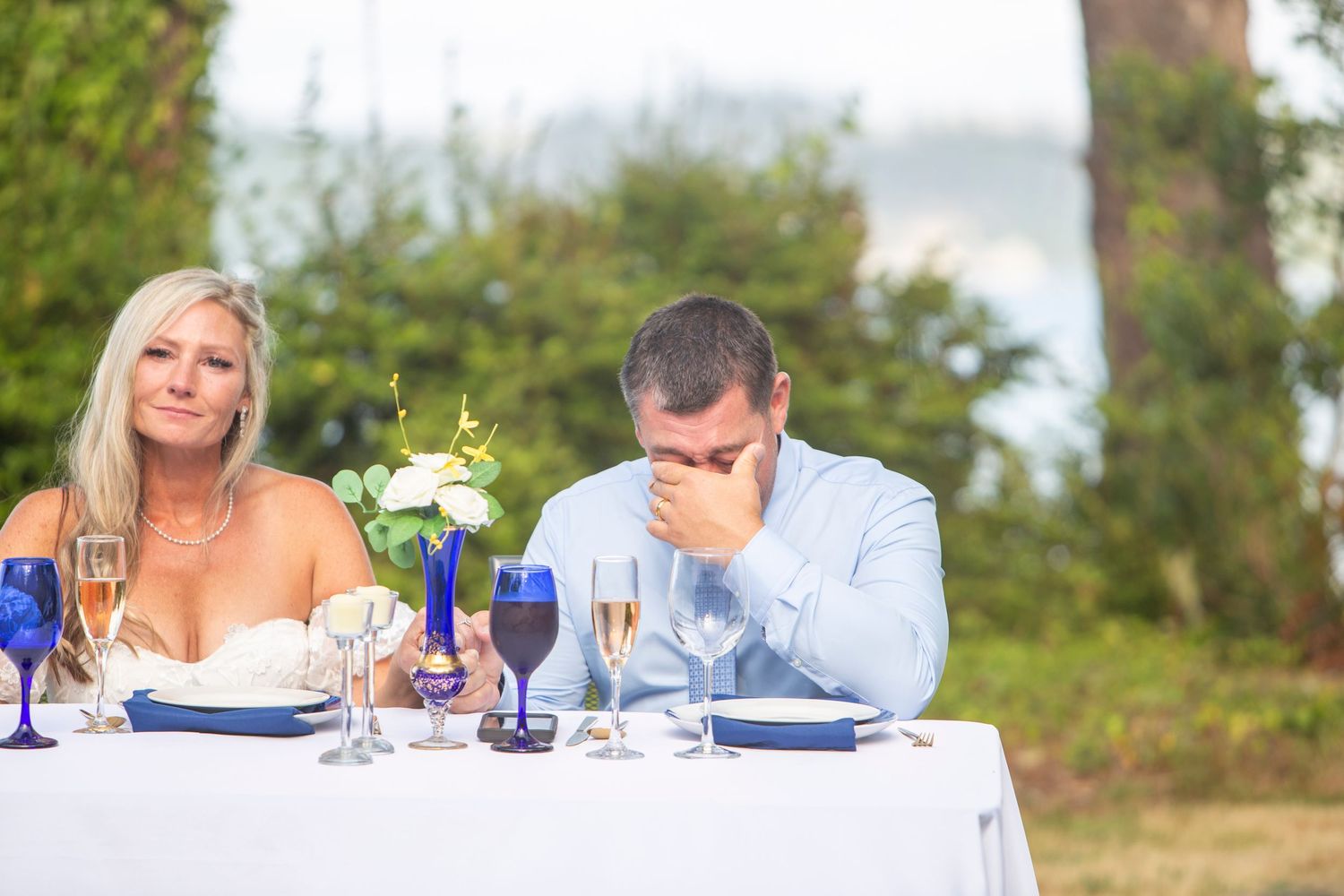Emotional groom covering face while bride smiles at outdoor wedding reception sweetheart table.