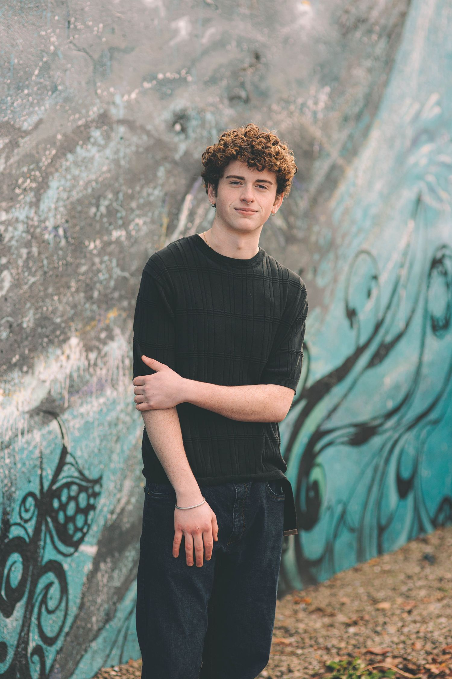 Young man with curly hair in black t-shirt leaning against blue graffiti mural wall.