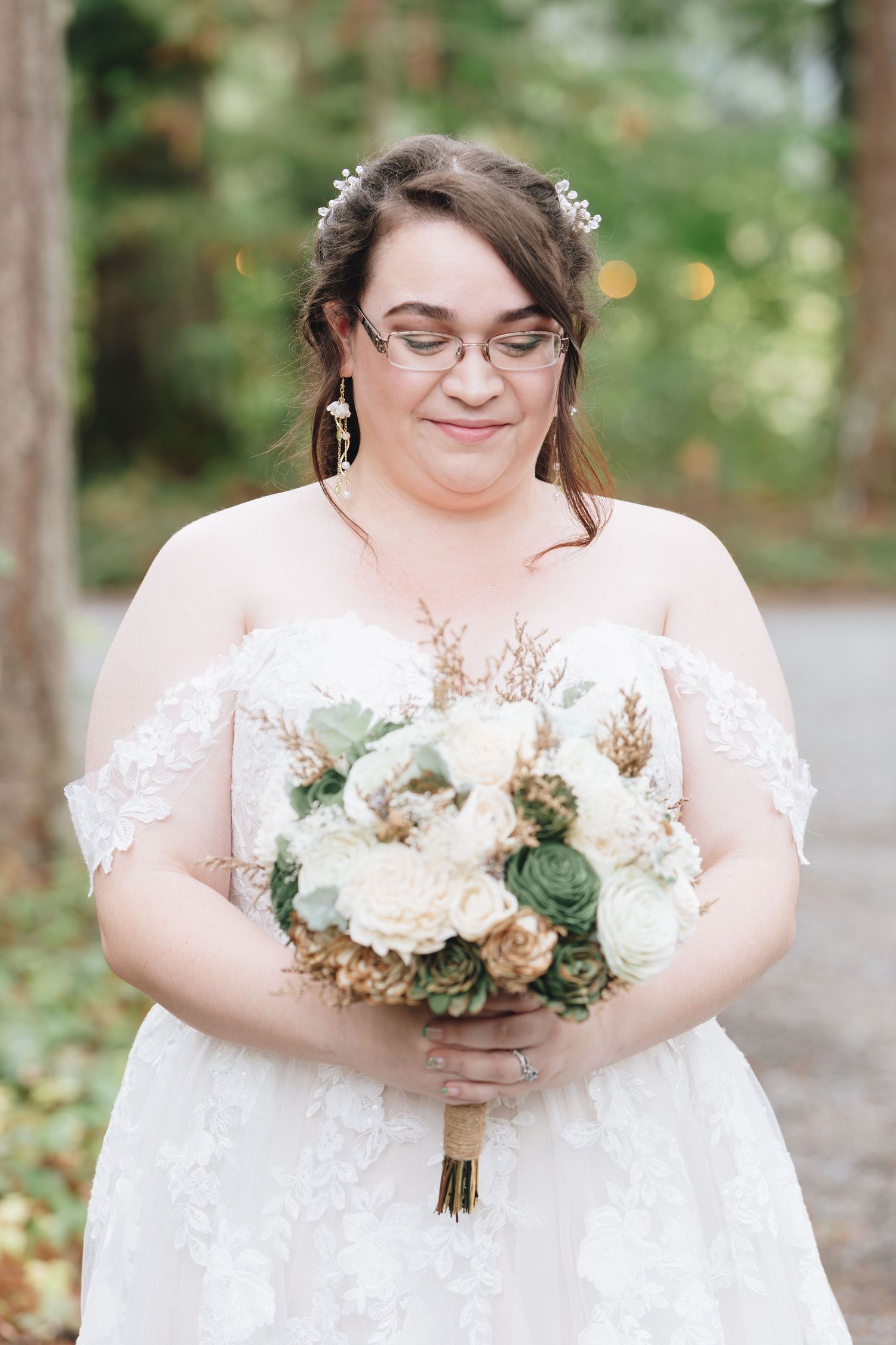 Bride in lace off-shoulder gown holding succulent and rose bouquet during outdoor forest wedding.
