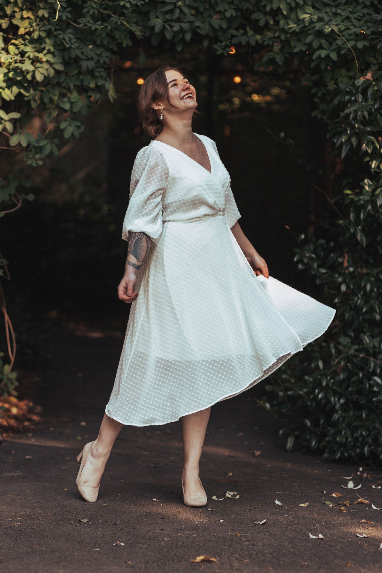 Joyful woman in white midi dress twirling near ivy-covered wall in outdoor setting.