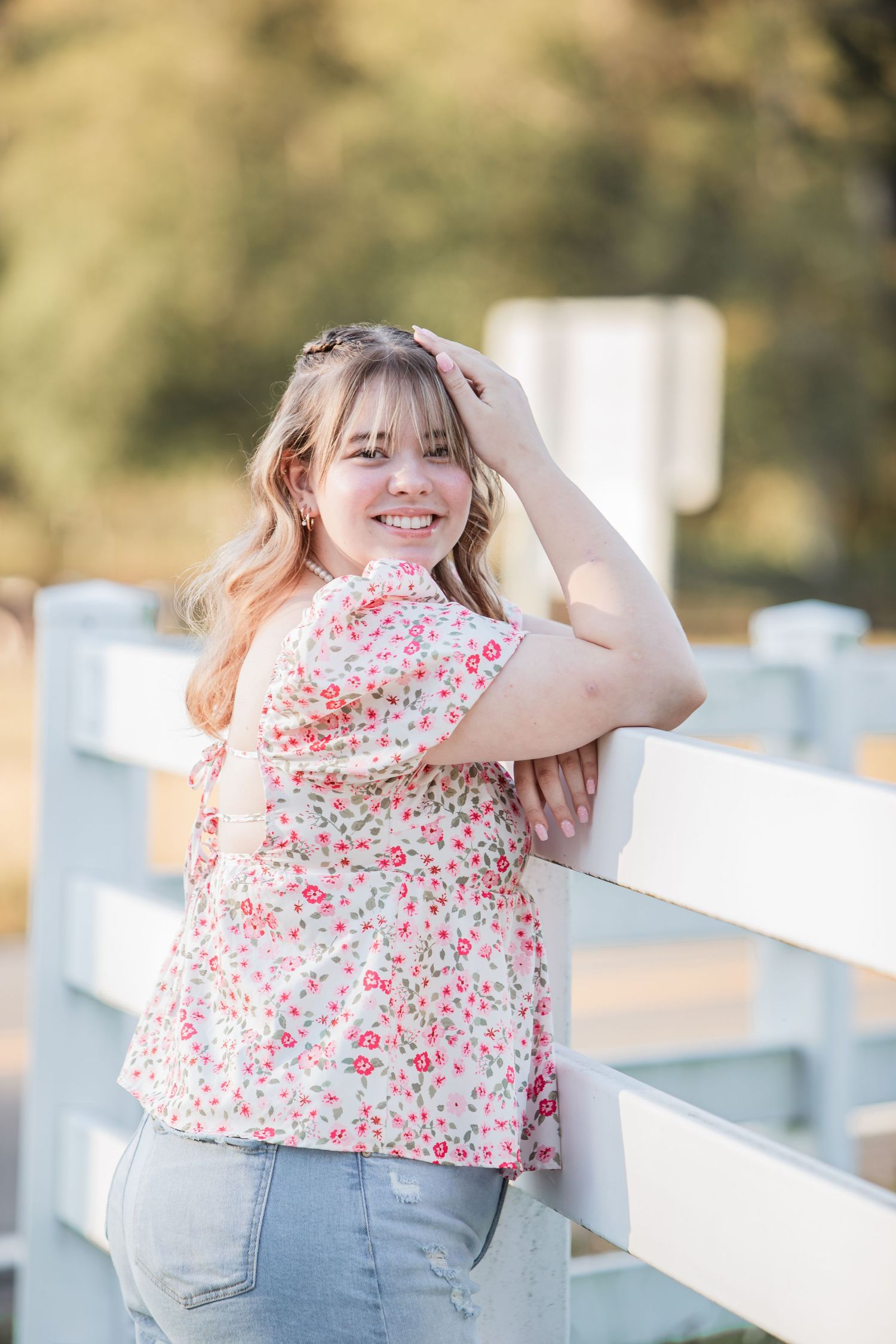 Smiling young woman in floral top leaning on a white fence in golden sunlight.