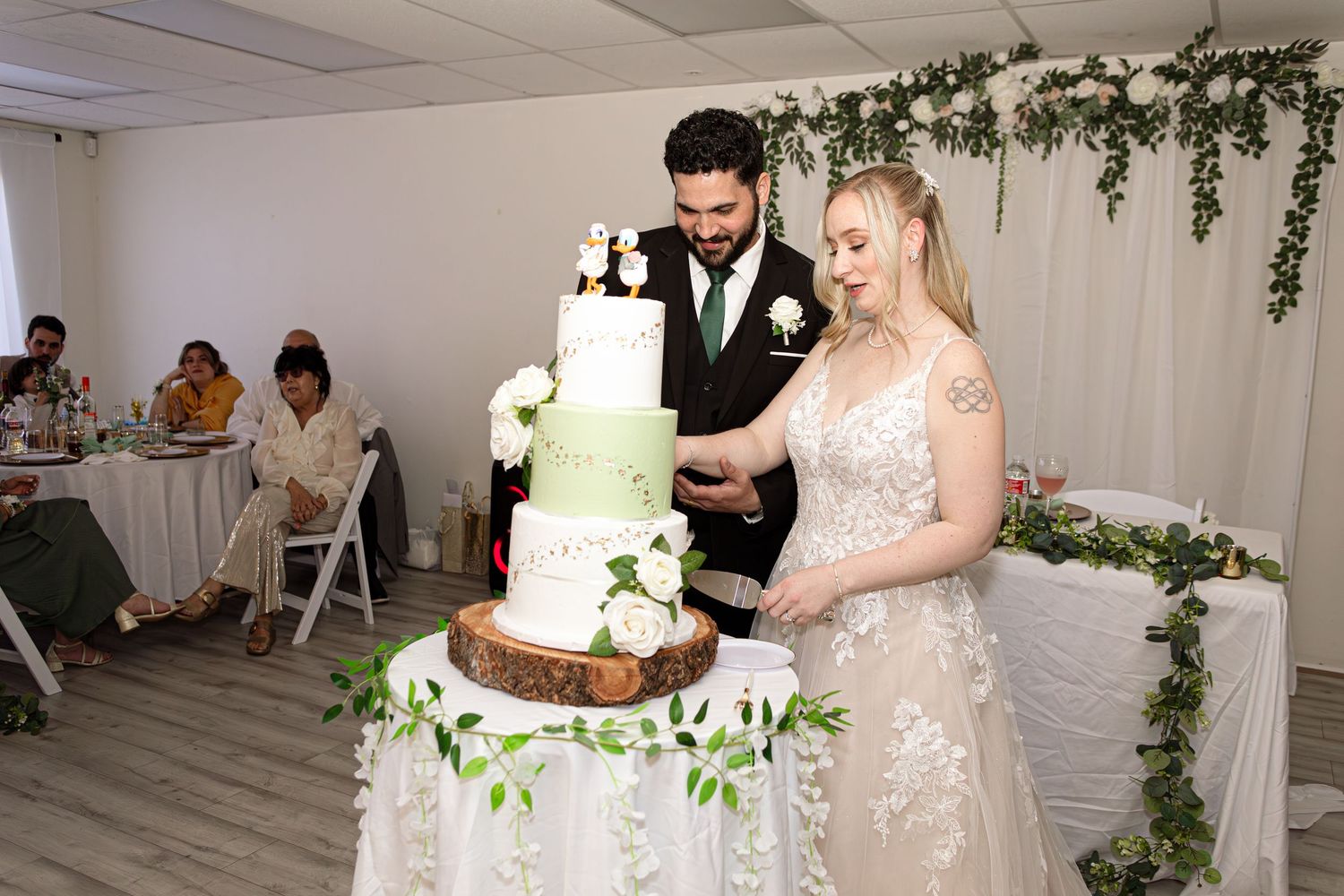 Bride and groom cutting a white four-tier wedding cake decorated with greenery and flowers at their reception.