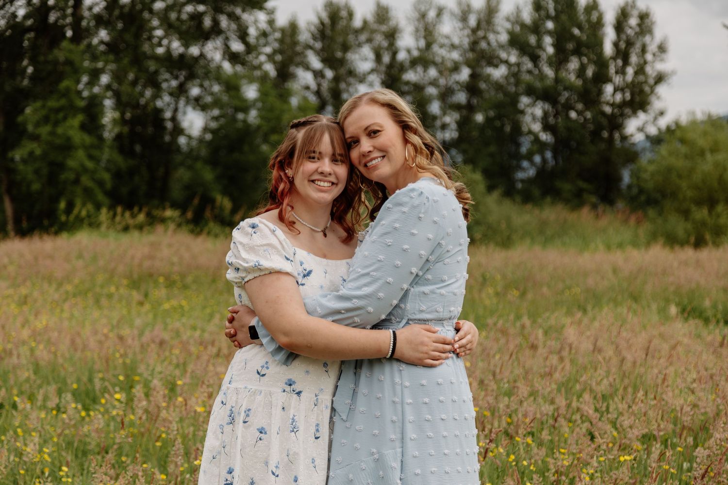 Two young women hugging and smiling together in a lush green meadow field outdoors.