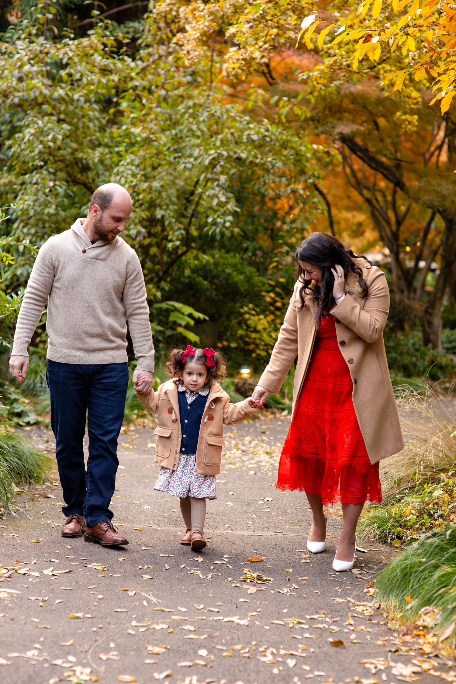 Parents walking with toddler daughter holding hands on a leaf-covered autumn garden path.