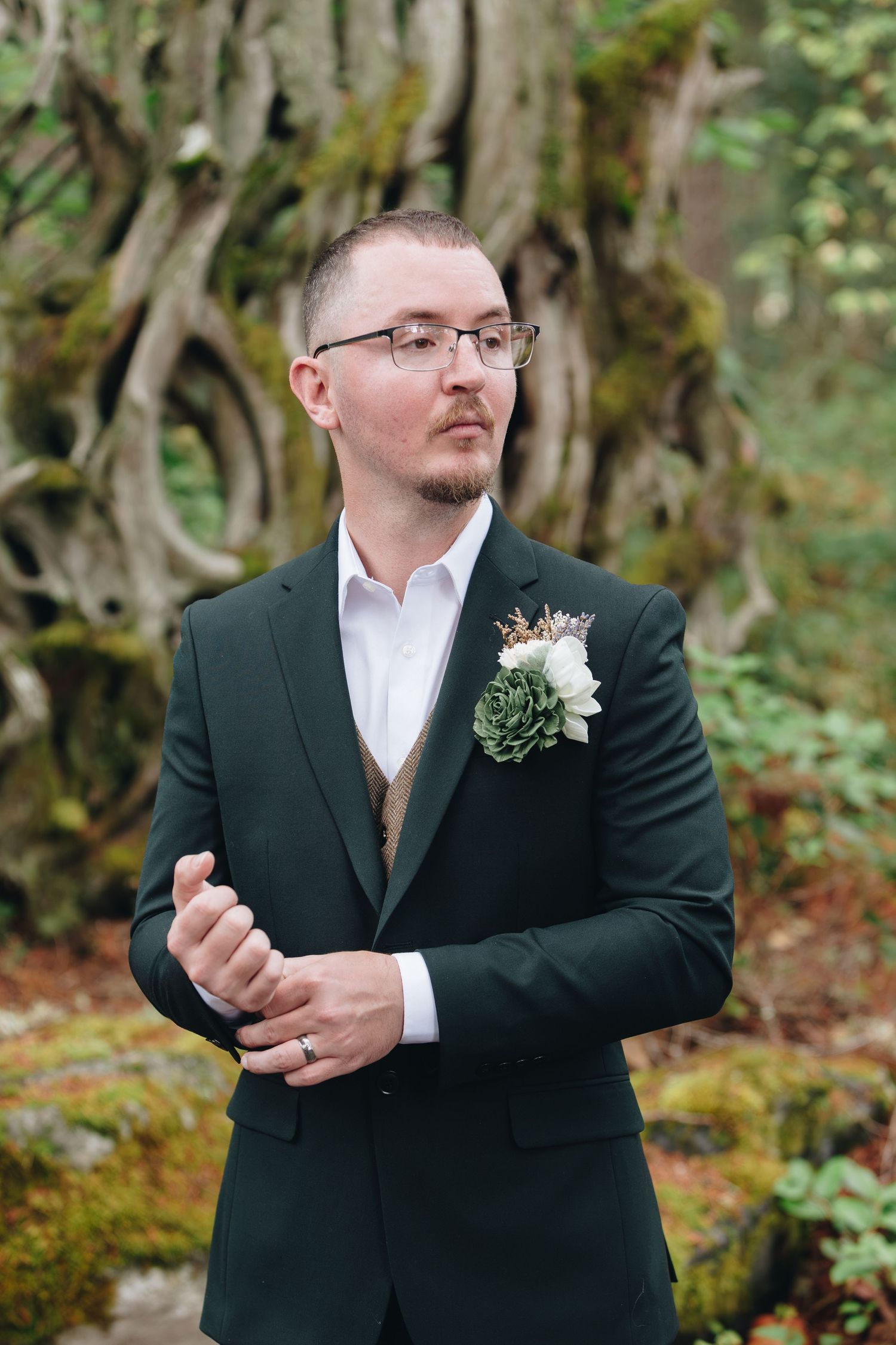 Groom in dark green suit with succulent boutonniere posing before moss-covered tree at wedding.