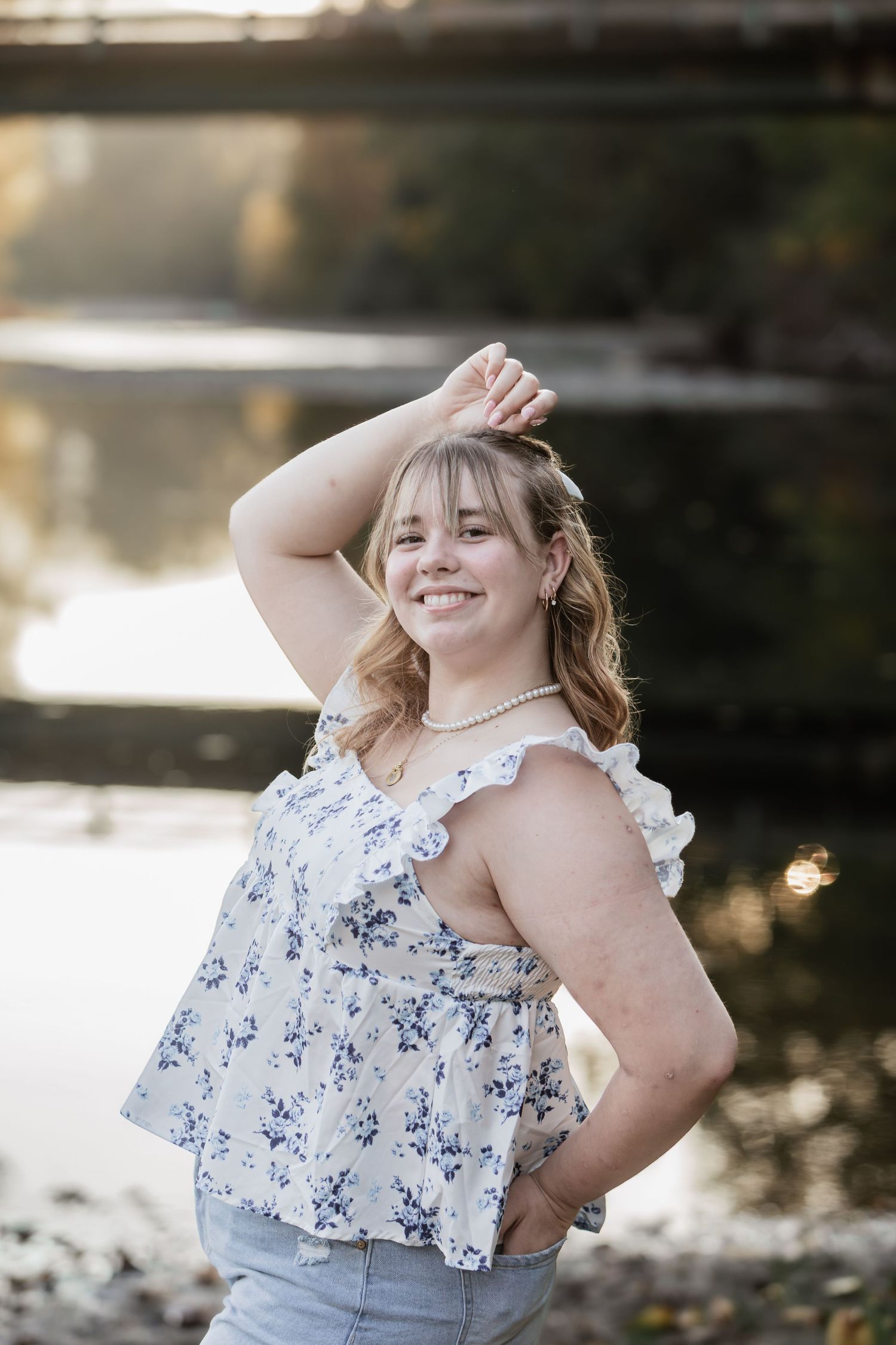 Smiling young woman in floral top posing by a reflective river with a bridge backdrop.