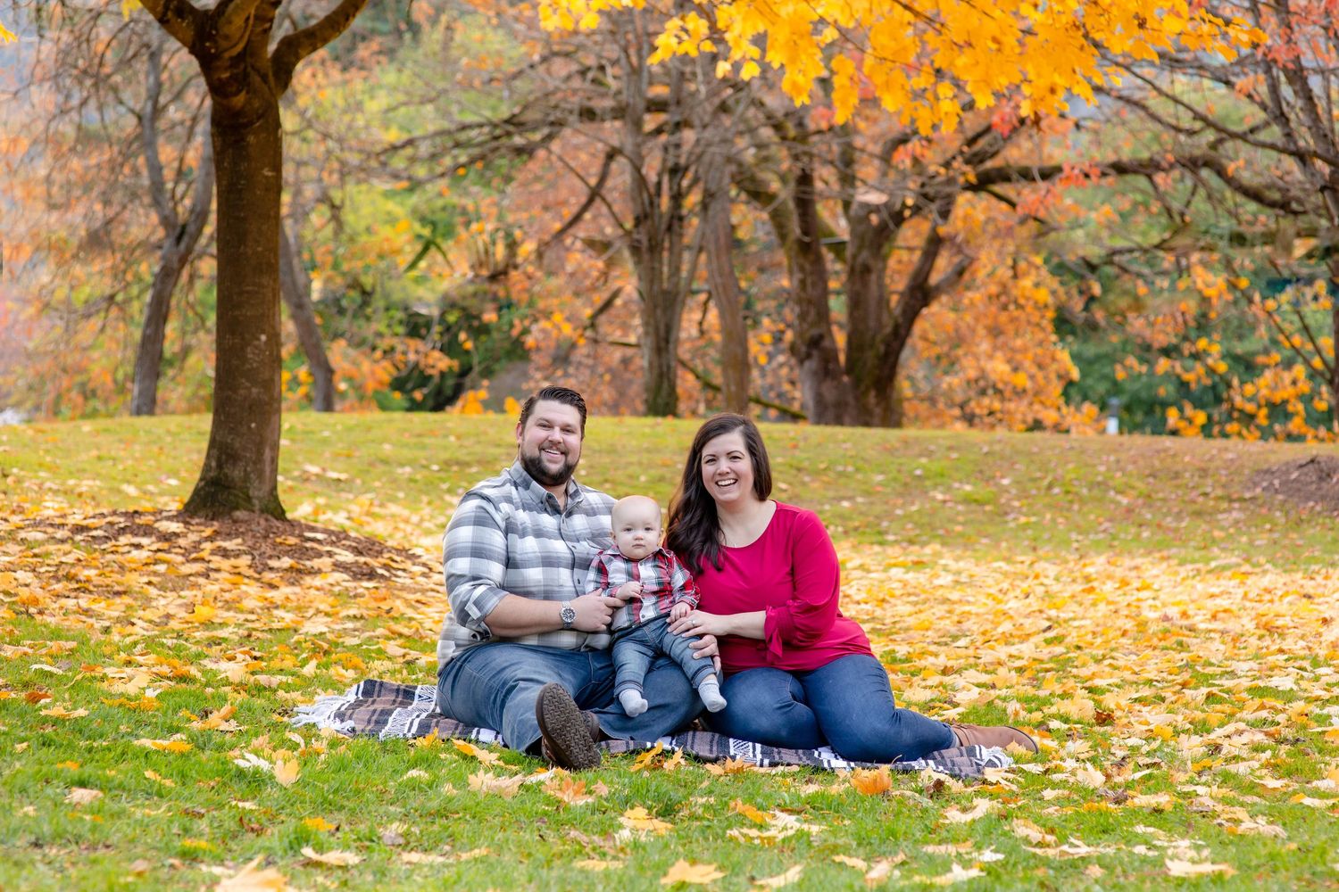 Young family with baby sitting on blanket surrounded by fallen autumn leaves in park.