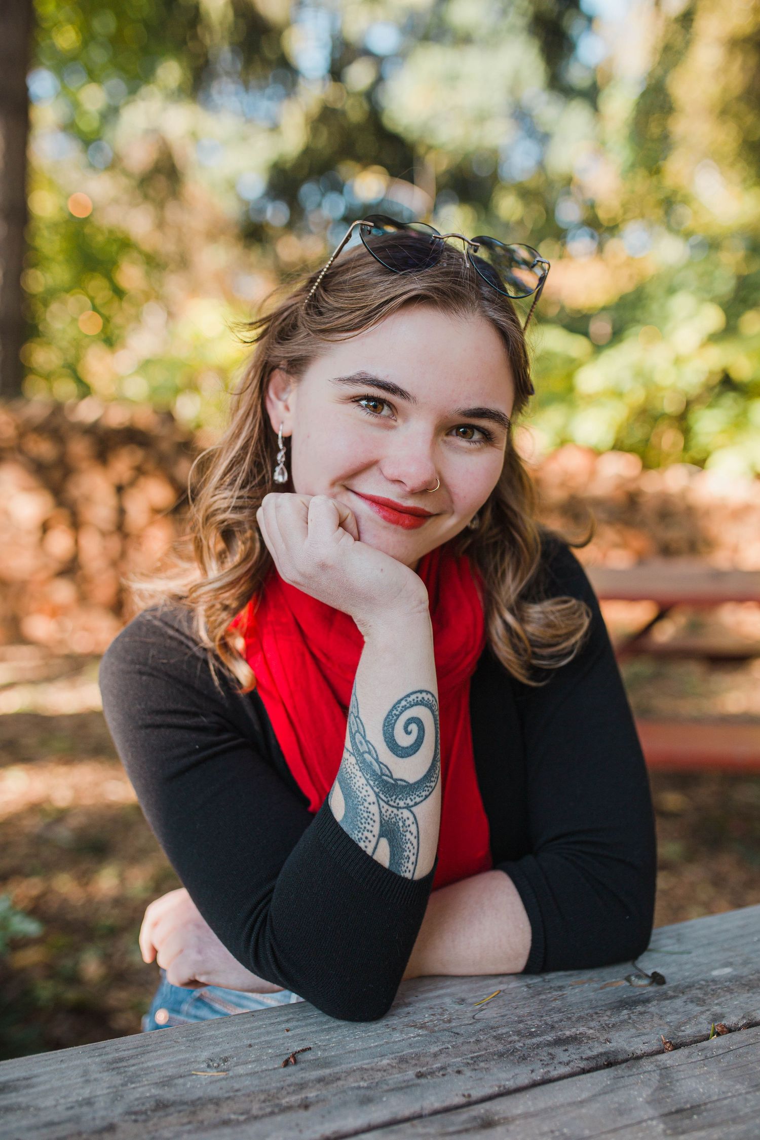 Smiling young woman with tattoo and red scarf resting chin on hand at outdoor table.