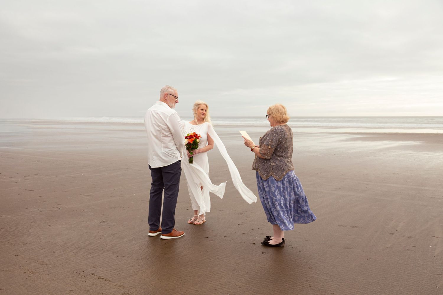 Couple exchanging vows on foggy beach with female officiant holding ceremony script.