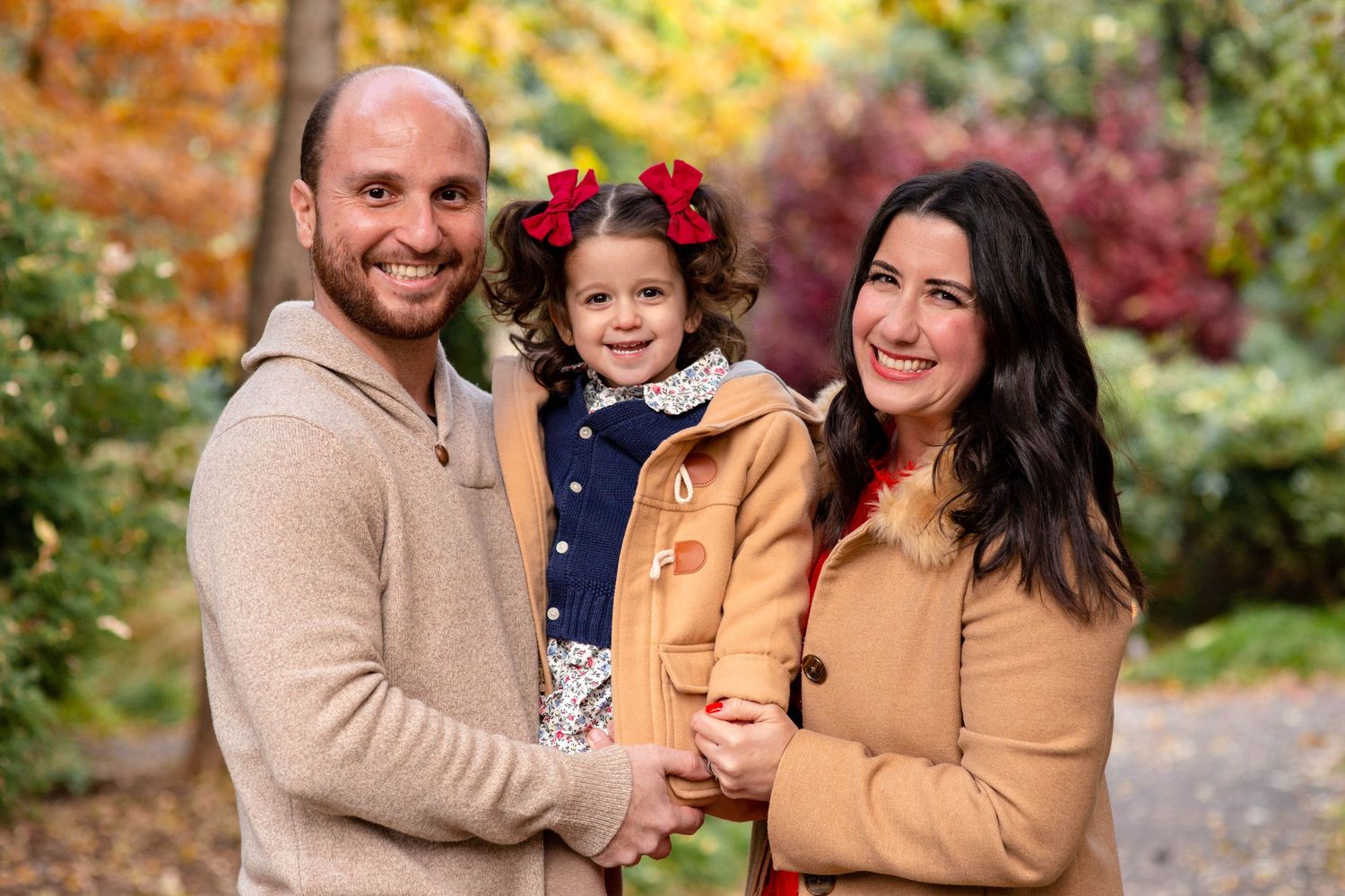 Happy family of three with a cute toddler girl in red bows posing together in a colorful fall park.