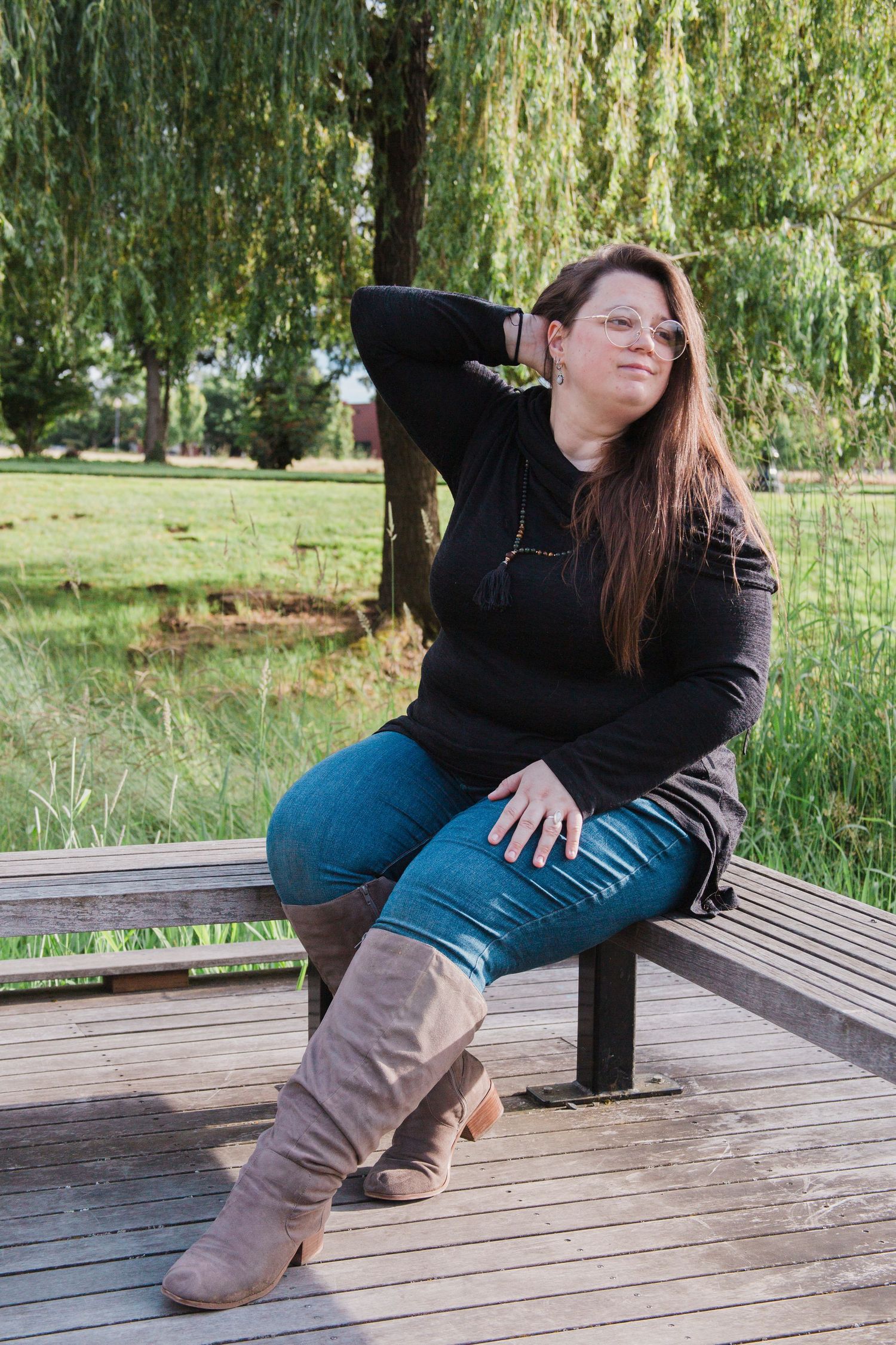 Woman in black top, jeans, and boots sitting on a wooden bench by a willow tree.