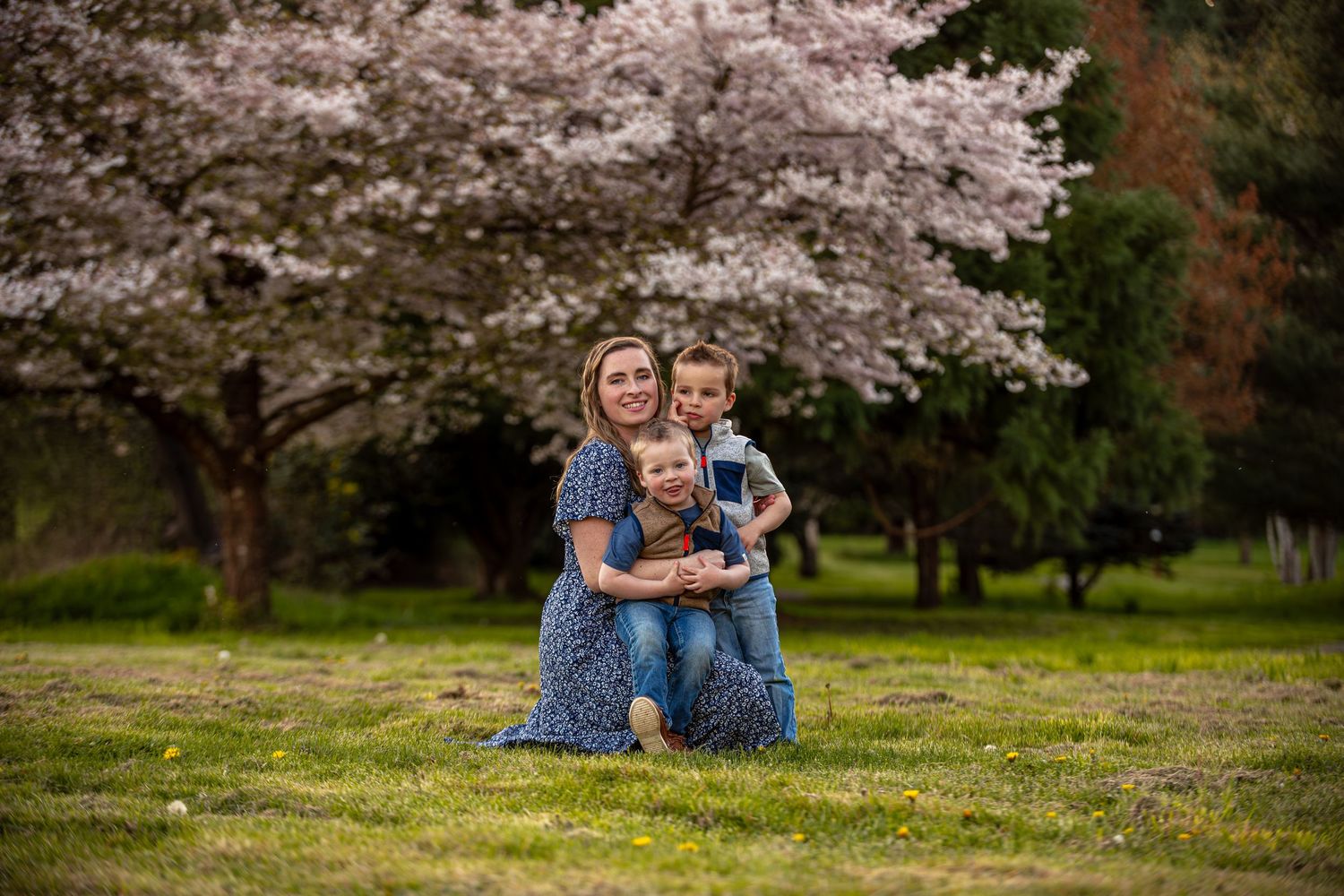 A mother sits on grass with two young children under a blooming cherry blossom tree in a lush green park.