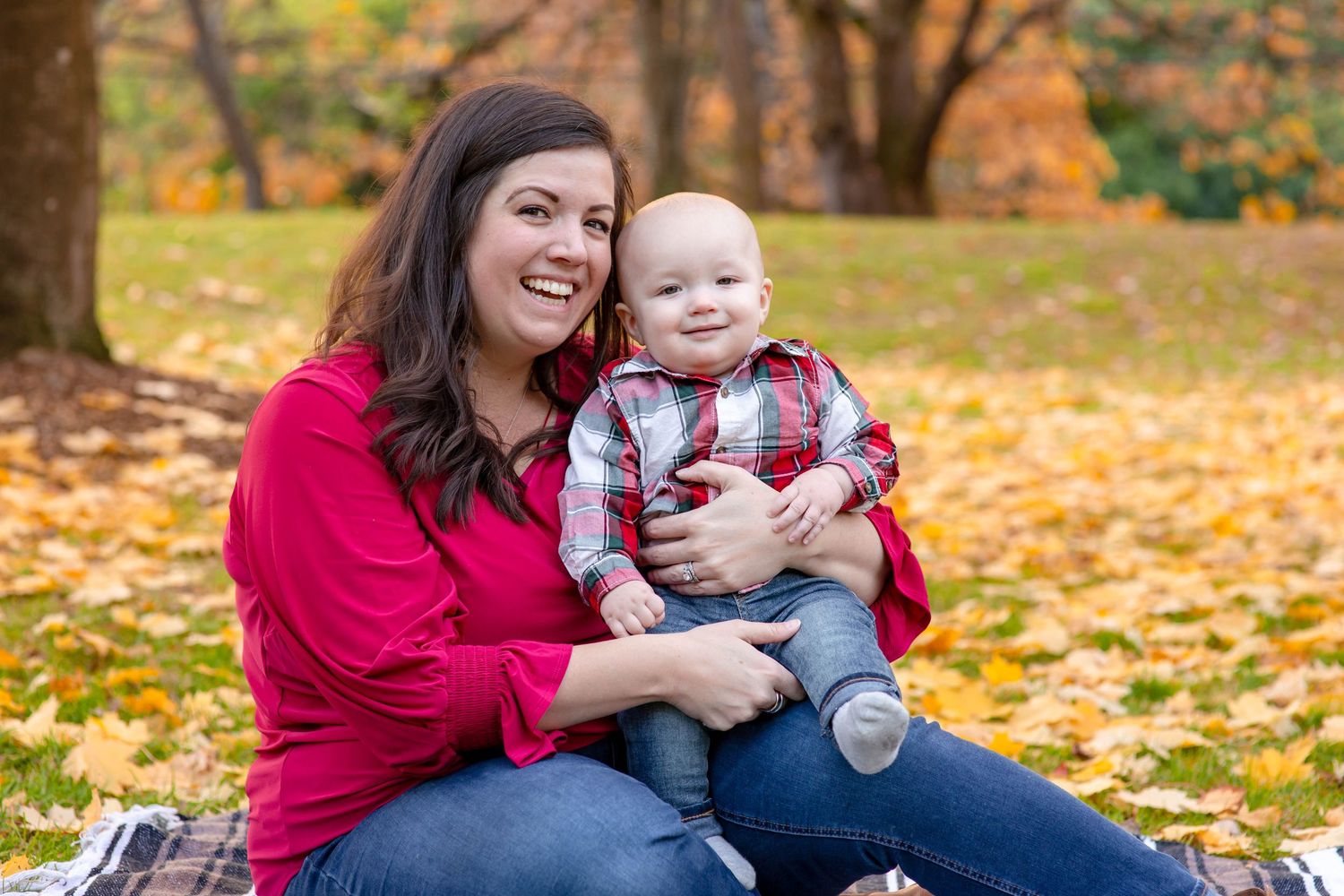 A smiling woman in red holds a happy baby in plaid shirt among autumn leaves in a park.