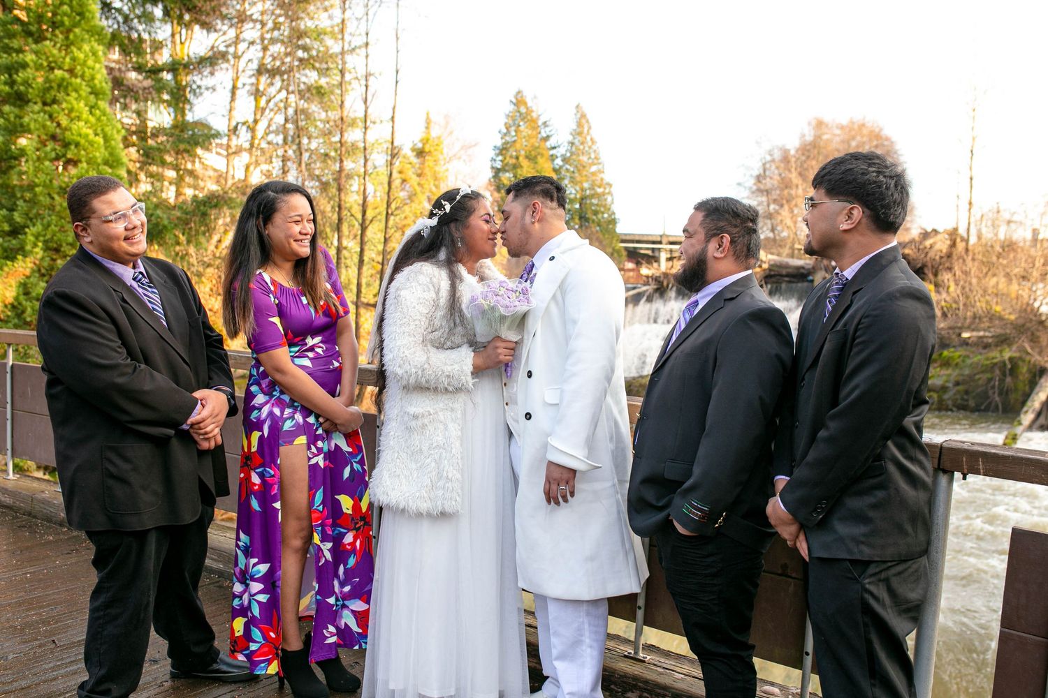 Bride and groom share a kiss surrounded by smiling wedding party guests on an outdoor bridge.
