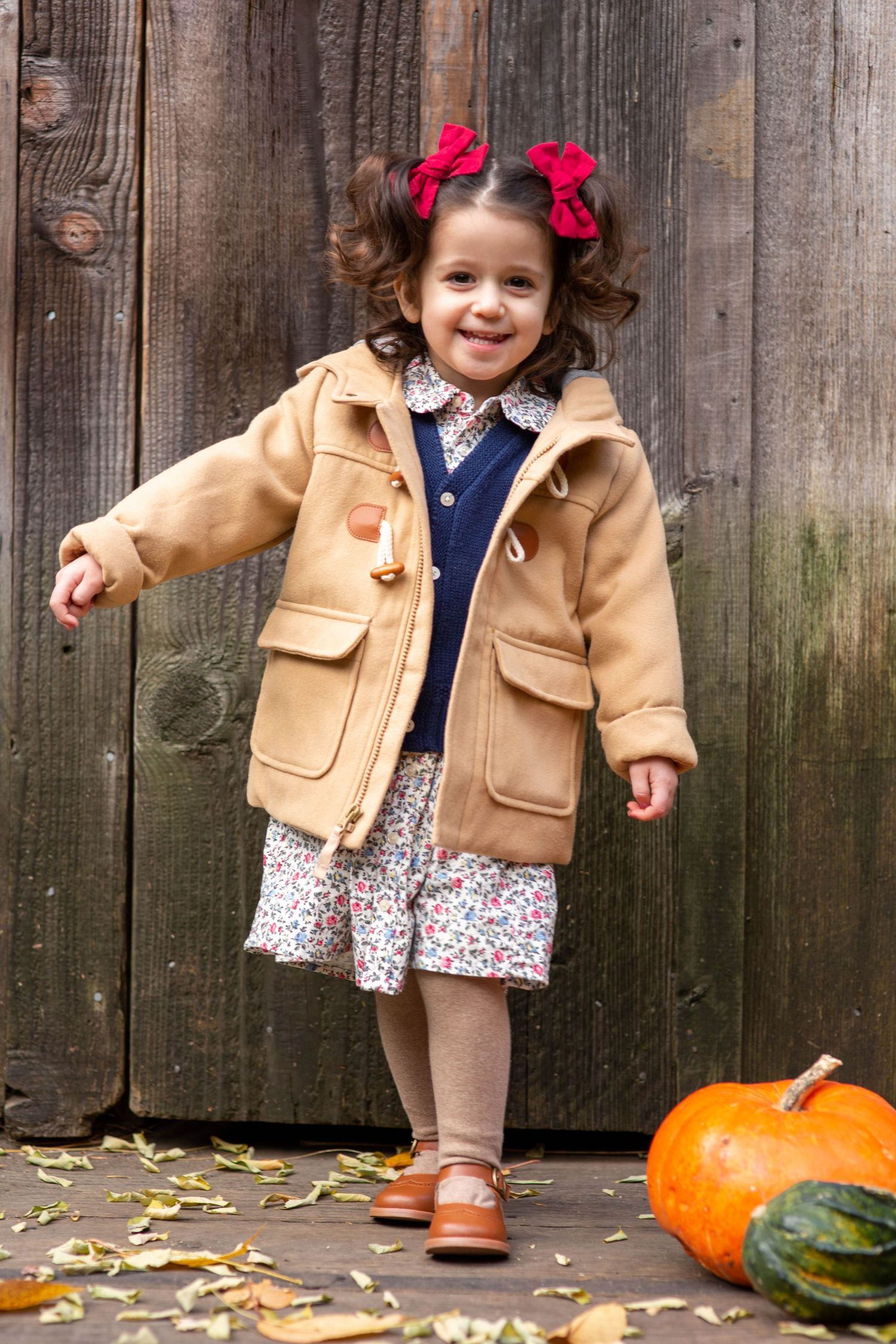 Adorable toddler girl in tan coat and red hair bows smiling beside a pumpkin in an autumn outdoor setting.