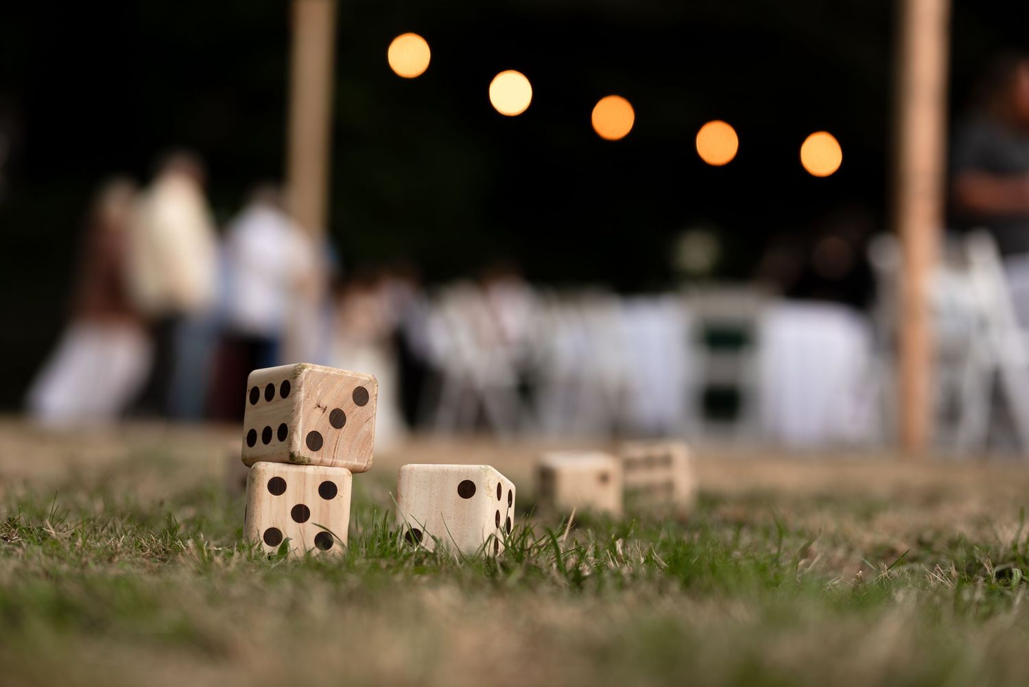 Large wooden lawn dice stacked on grass at an outdoor evening wedding reception with string lights.