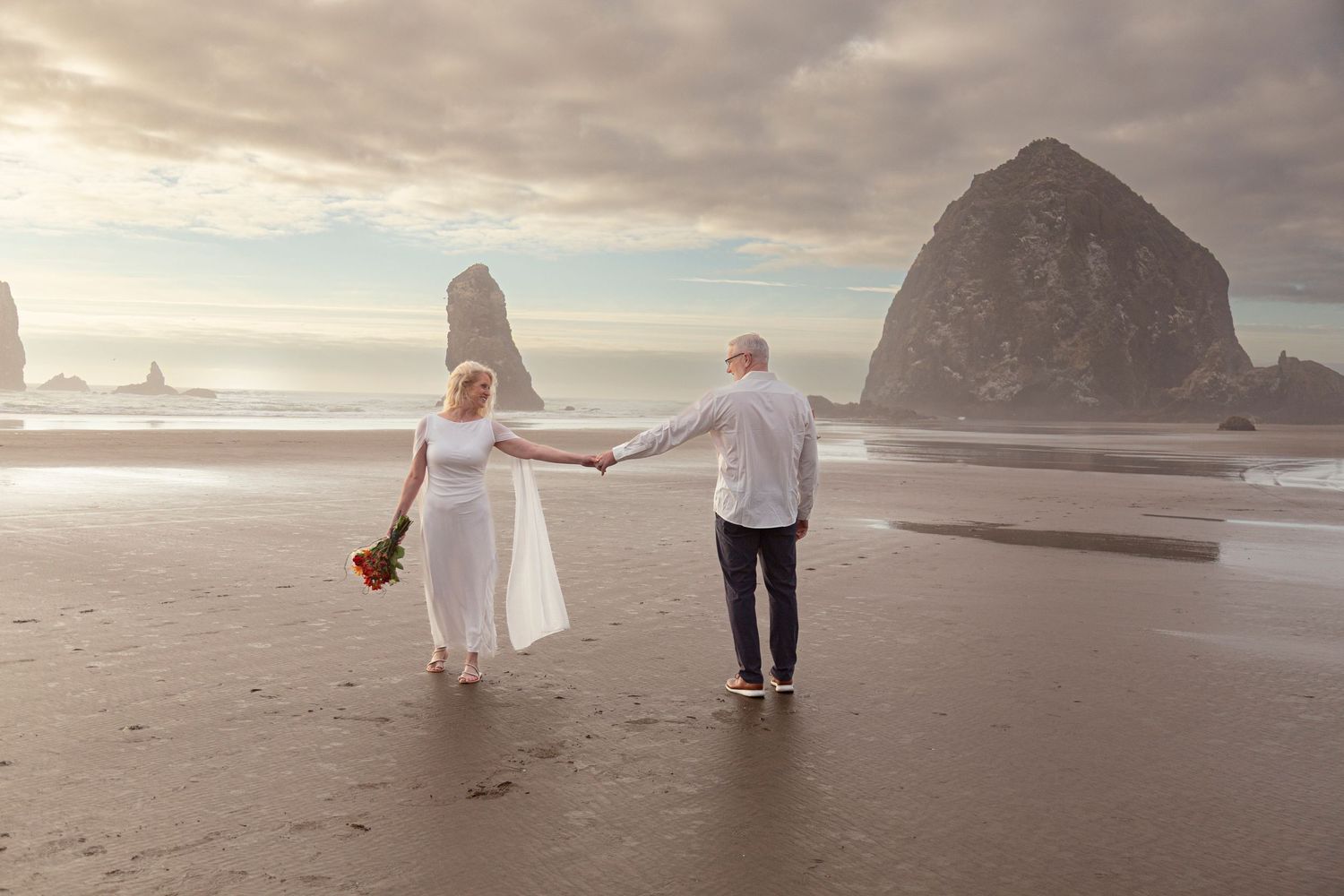Couple holding hands on Cannon Beach with Haystack Rock in background during beach wedding.