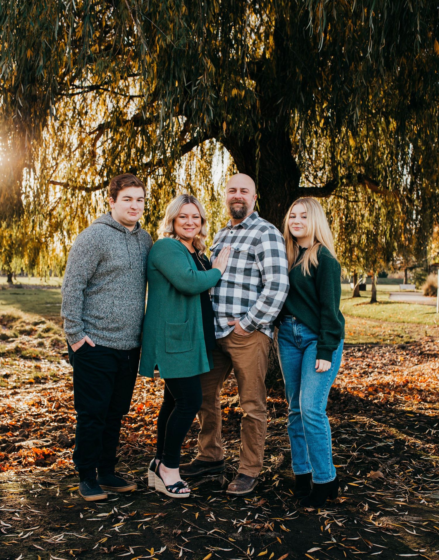 Family of four posing under a willow tree in autumn park, wearing green and plaid outfits.