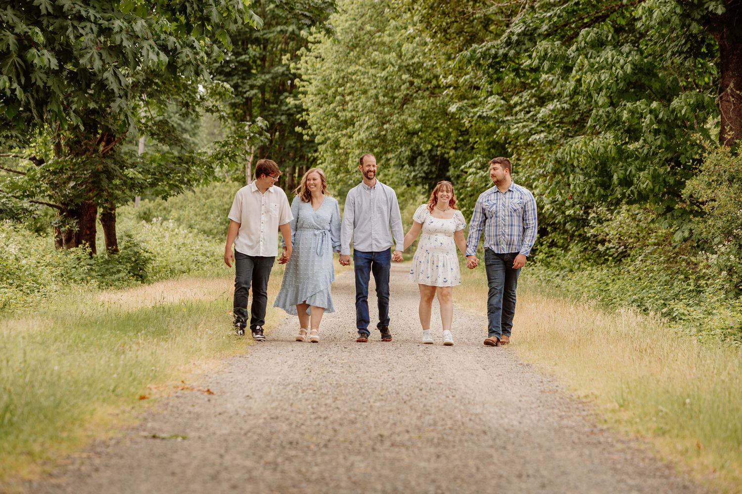 Family of five walking hand in hand down a tree-lined gravel path in a lush summer forest.