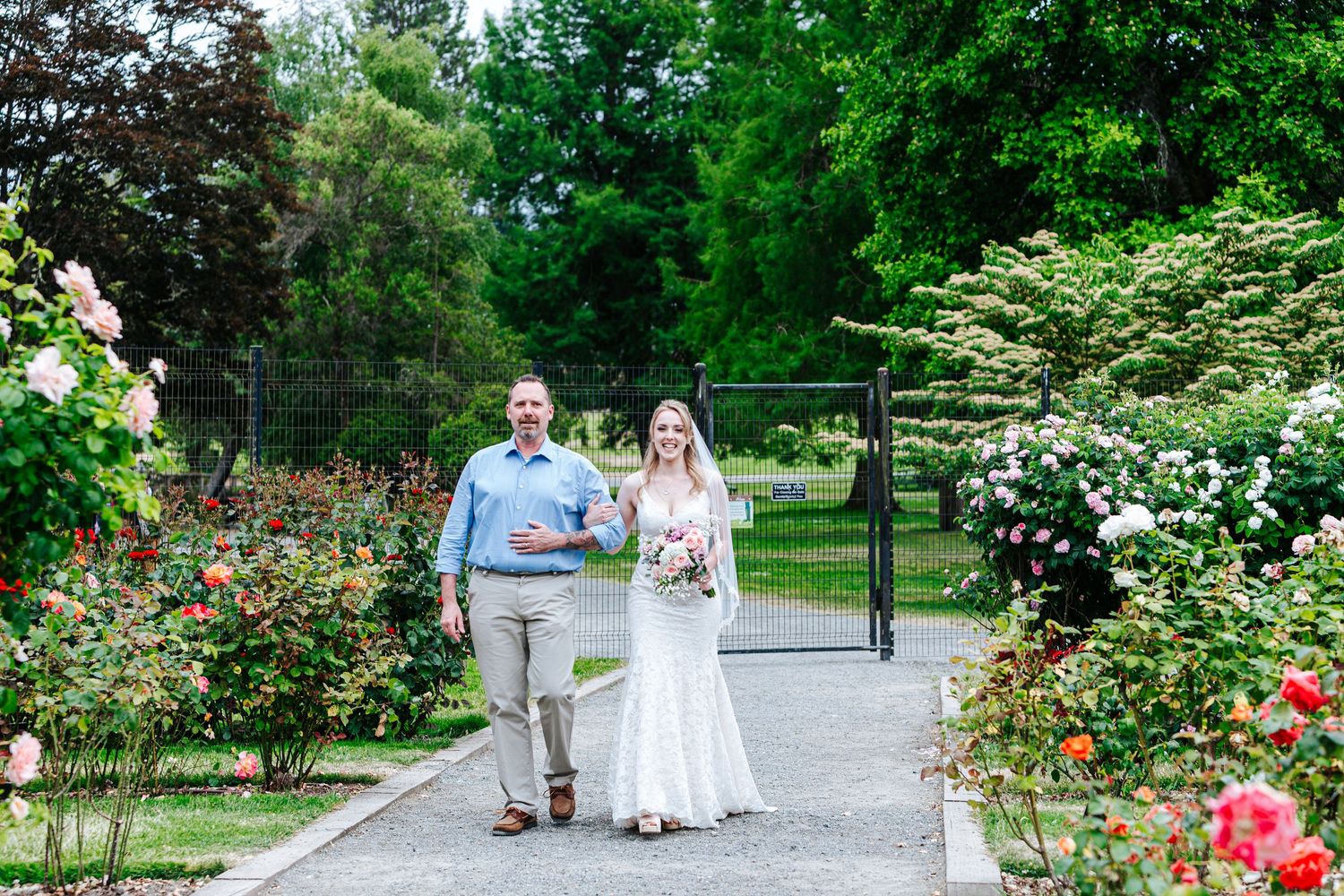 Bride in lace gown walks with escort through rose garden on wedding day.