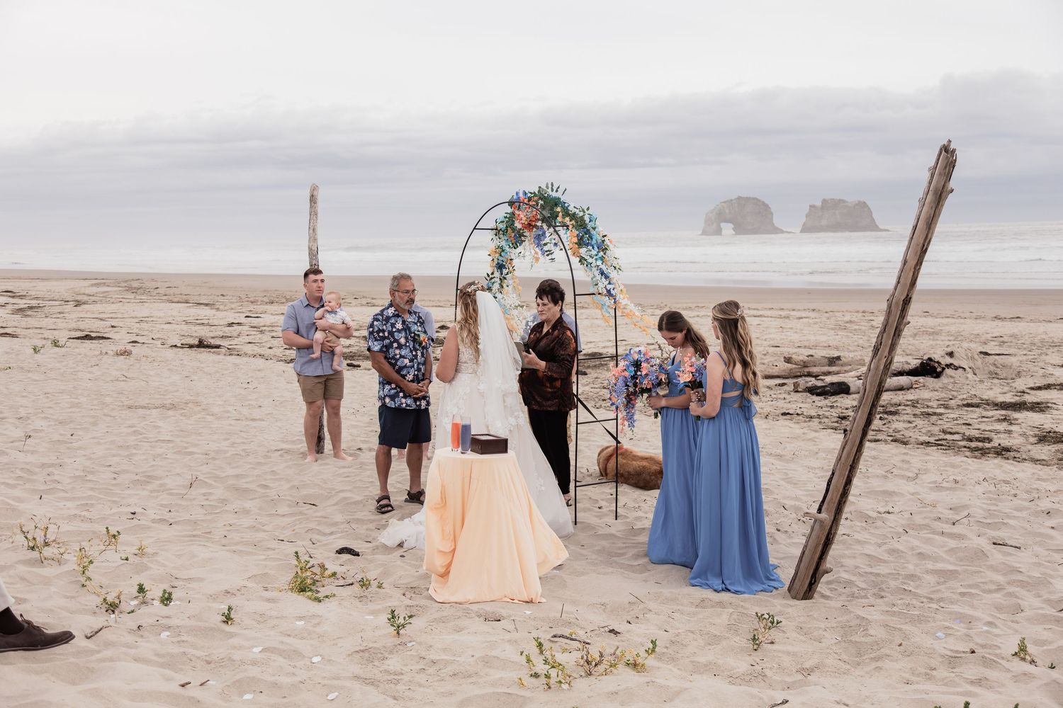 Beach wedding ceremony at arch with bridesmaids in blue dresses on overcast day.