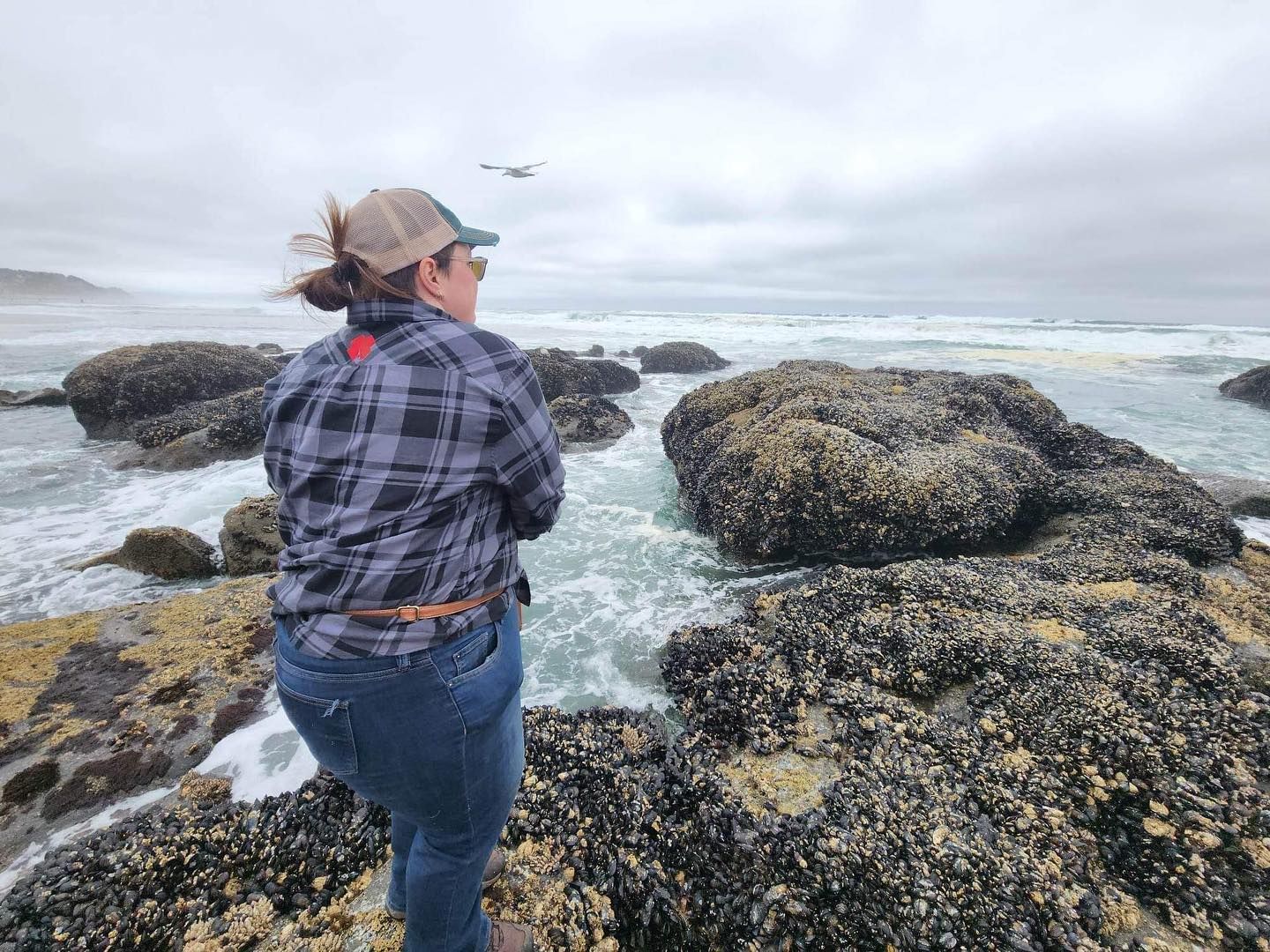 Woman in flannel shirt standing on rocky coastal shoreline watching ocean waves.
