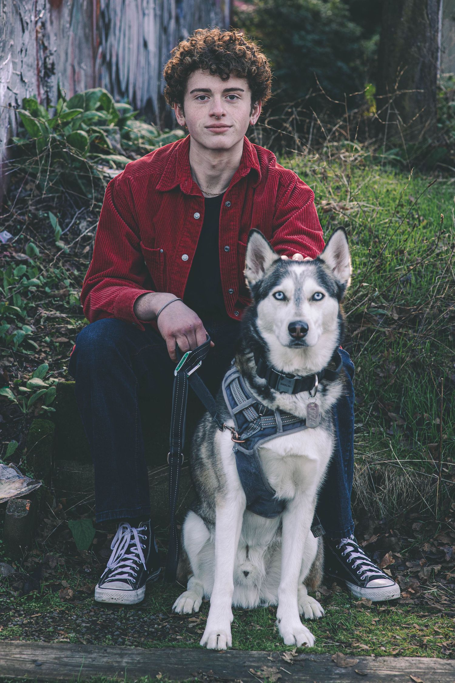 Young man in red jacket sitting with Siberian Husky dog in outdoor garden setting.