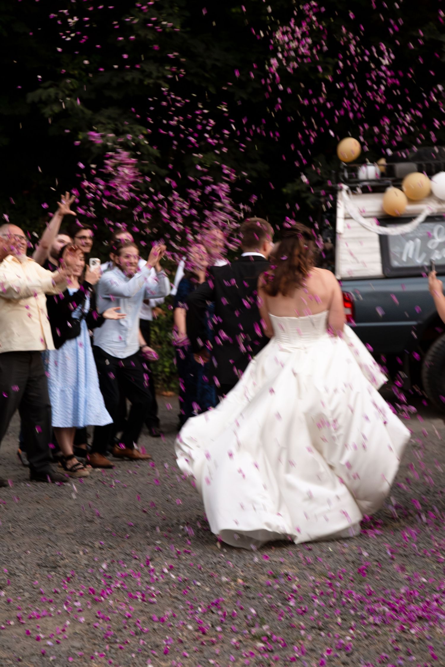 Bride in white gown walks through shower of pink confetti as wedding guests celebrate outdoors.