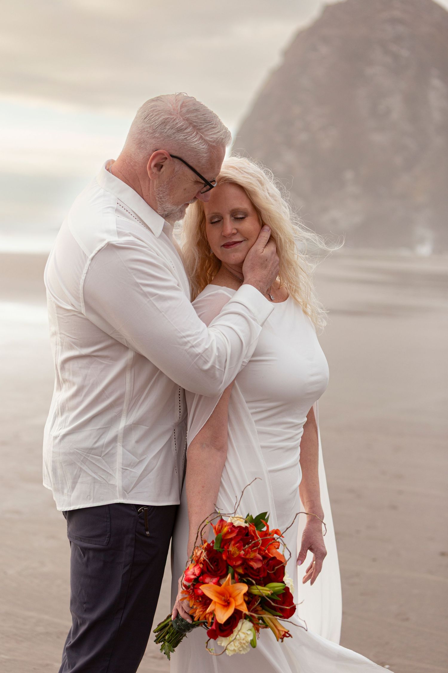 Couple in white attire share romantic beach moment, woman holds vibrant orange bouquet near iconic Haystack Rock.
