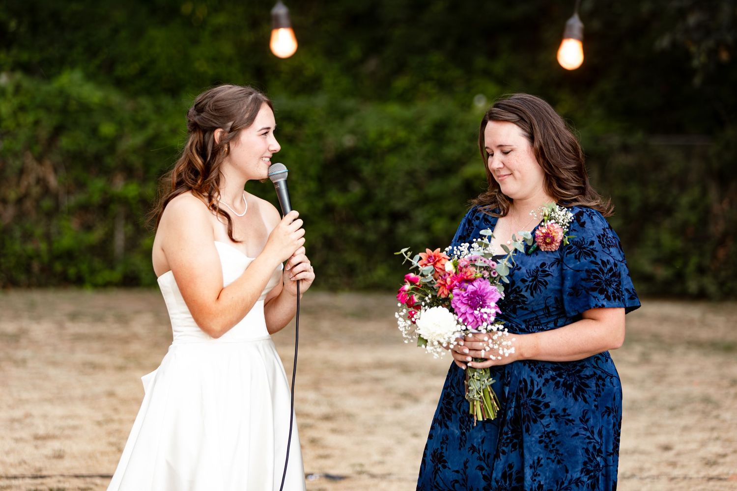 Bride in white dress speaks into microphone beside woman in blue floral dress holding colorful bouquet at outdoor wedding.