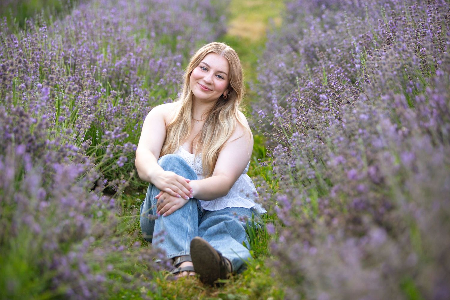 Young blonde woman sitting cross-legged in a vibrant purple lavender field, smiling warmly at the camera.