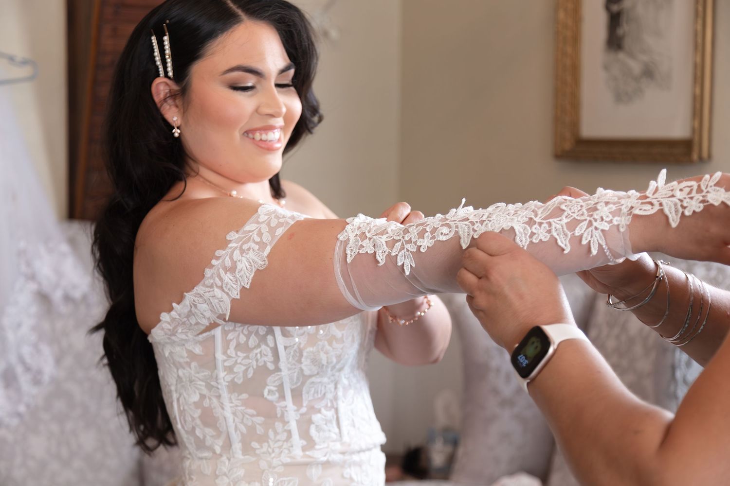 Smiling dark-haired bride in lace wedding dress being helped with her sleeves while getting ready.