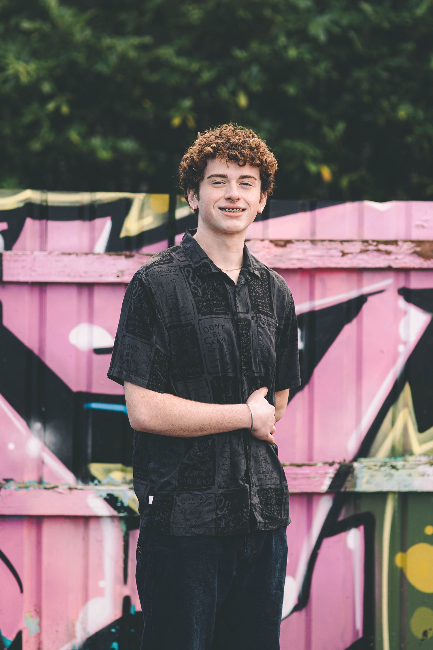Smiling teen boy with curly hair in black shirt standing before colorful graffiti wall.