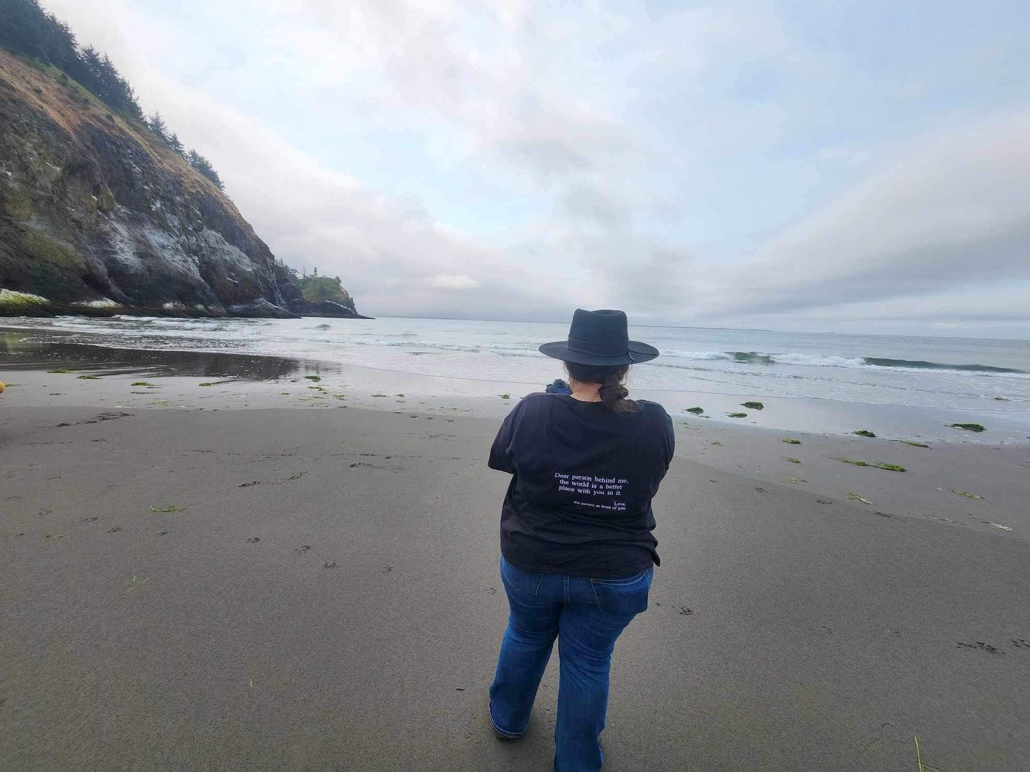 Person in a hat standing on a sandy beach with rocky cliffs and ocean waves.