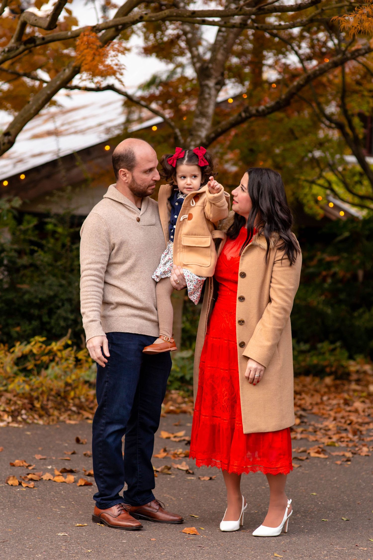 Family of three posing outdoors in fall, parents holding toddler girl with red bow, surrounded by autumn foliage.