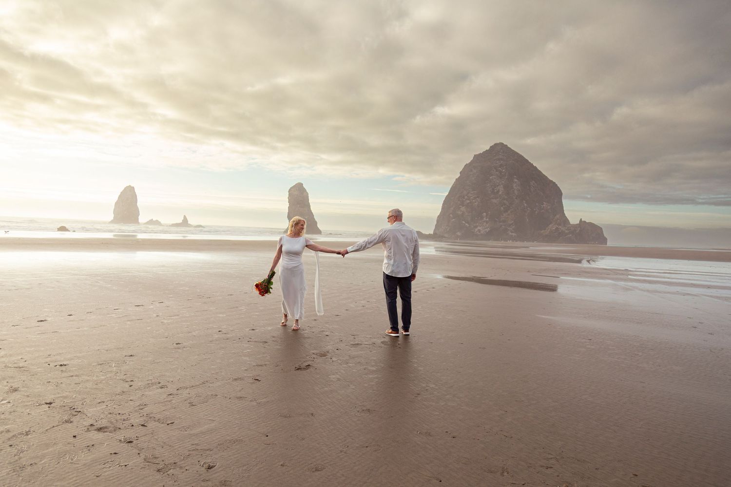 Couple holding hands walking on Cannon Beach with Haystack Rock backdrop on their wedding day.