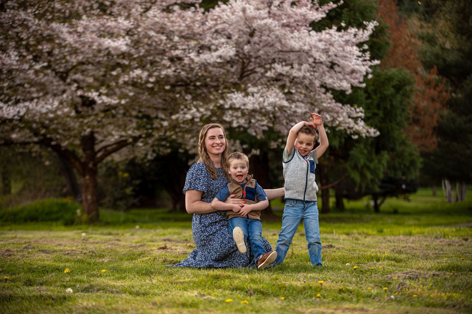 Smiling mother with two young boys playing on grass beneath blooming cherry blossom trees in spring.