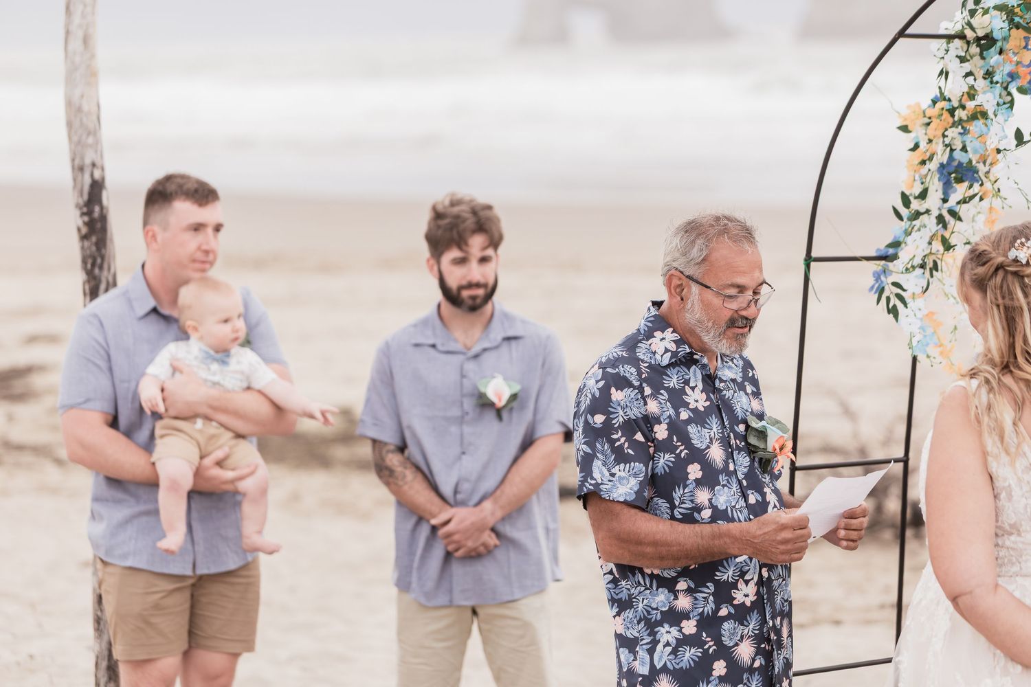 Groomsmen smiling during beach wedding ceremony as officiant reads vows outdoors.