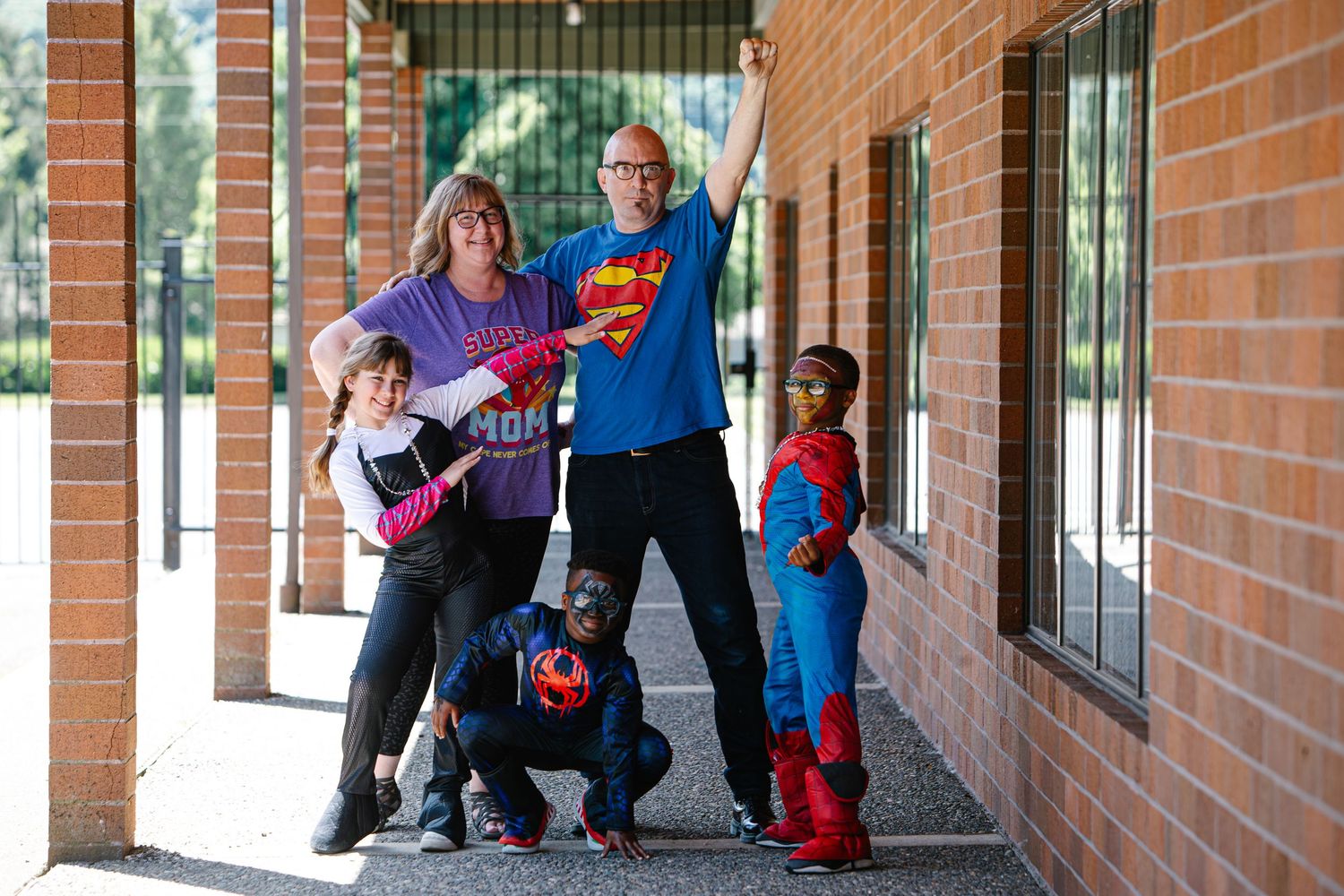 Family dressed as superheroes poses outside brick building, including Superman and Spider-Man costumes.