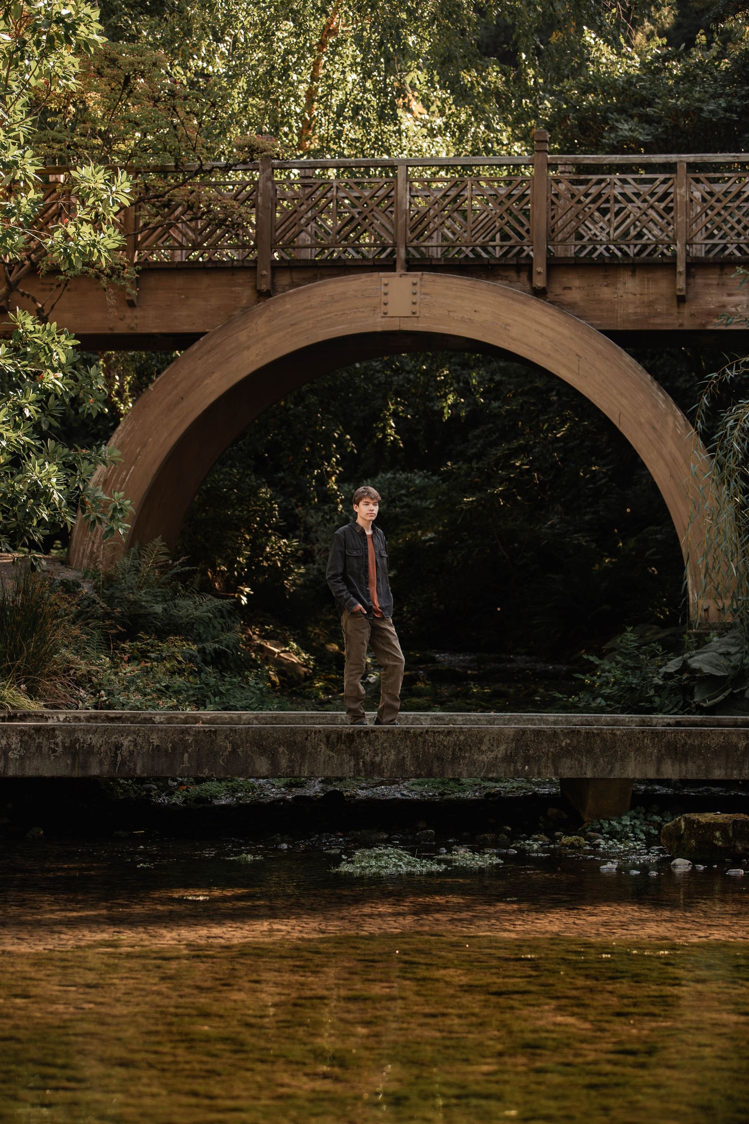 Young man standing on a stone path beneath a large arched bridge surrounded by lush greenery near a tranquil pond.