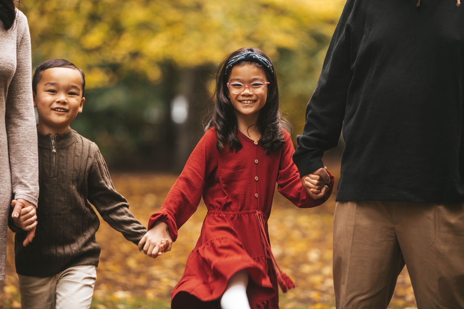 Two young Asian children holding hands with parents while walking through autumn park.
