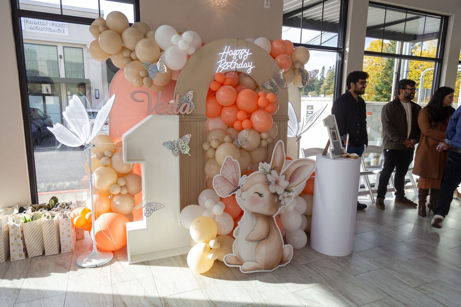 First birthday party decoration with balloon arch, bunny cutout, number 1, and Happy Birthday sign in peach and white tones.