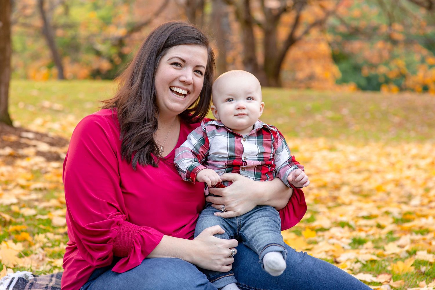 Smiling mother holding happy baby boy sitting together on autumn leaves in a park.