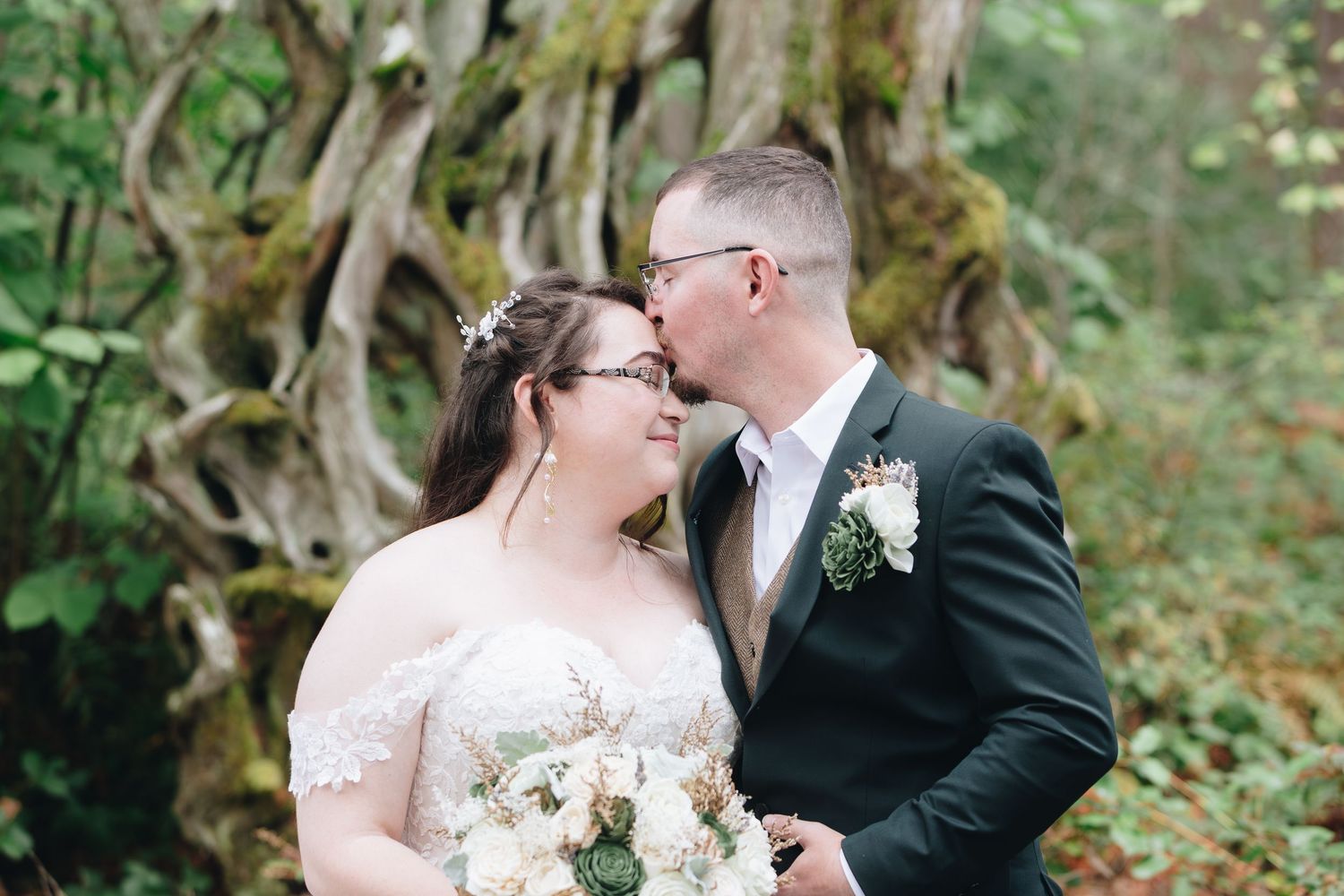 Bride and groom sharing a tender kiss near a mossy tree during their forest wedding.