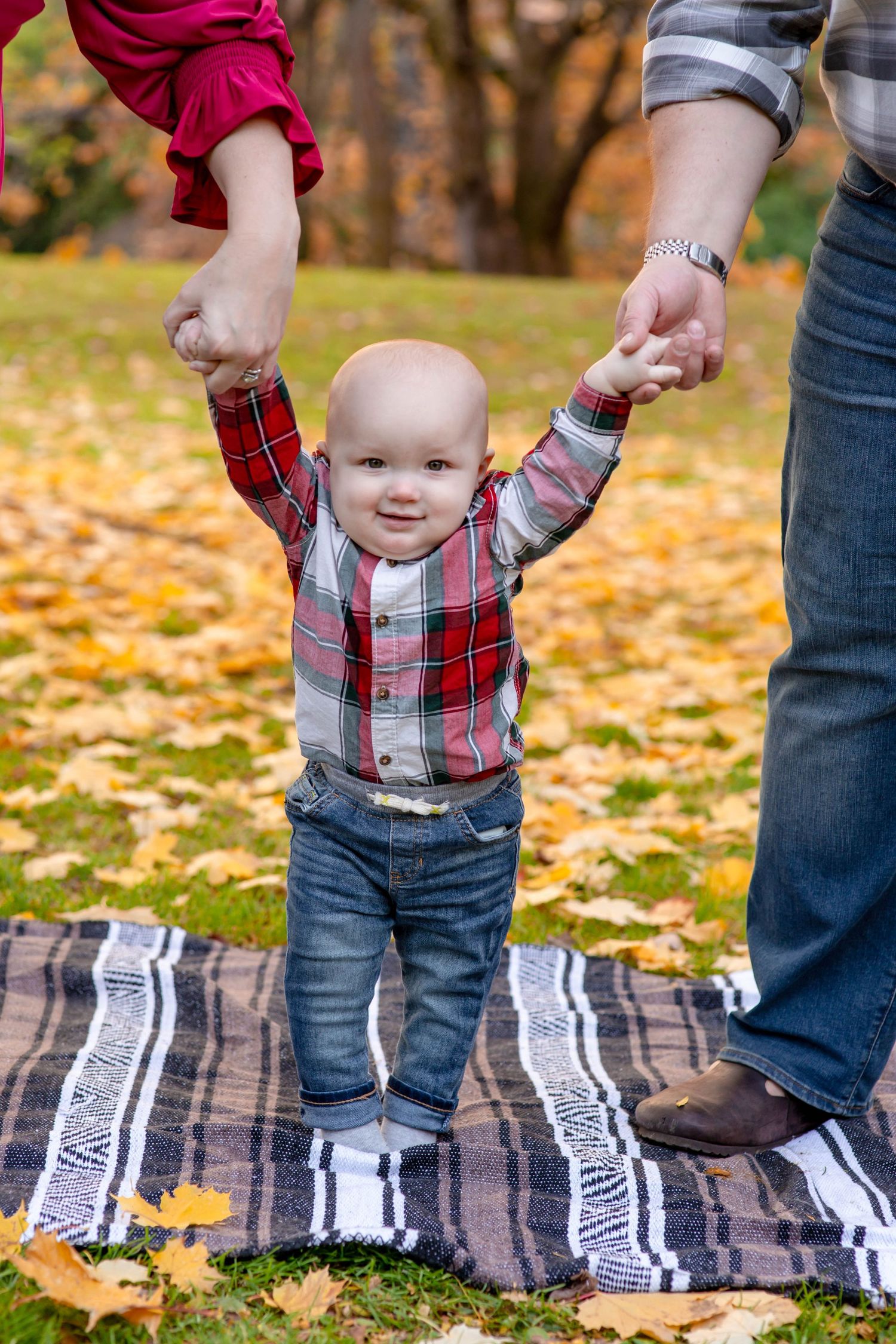 Happy baby in plaid shirt taking first steps while holding parents hands on autumn blanket.