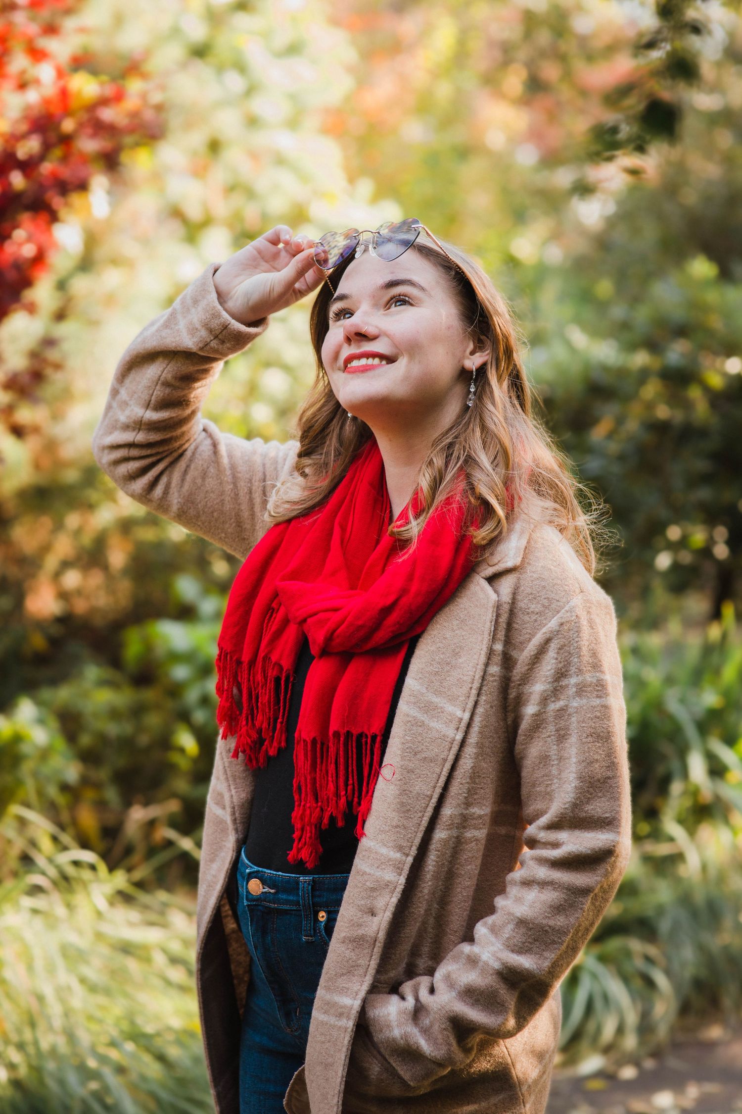 Happy young woman in tan blazer and red scarf holding sunglasses in autumn garden.