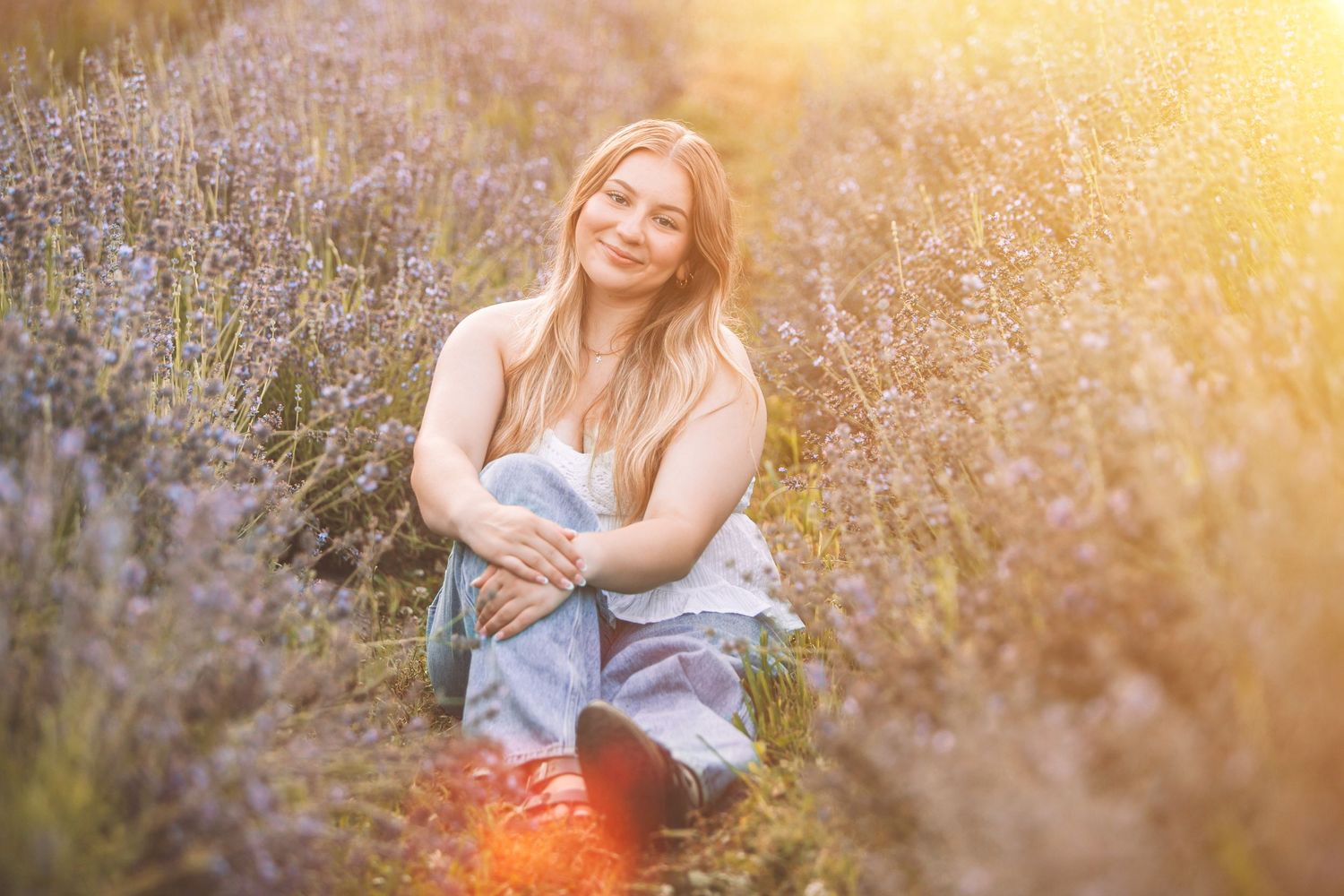 Young woman sitting in a lavender field, smiling warmly in golden sunlight, wearing a white top and jeans.