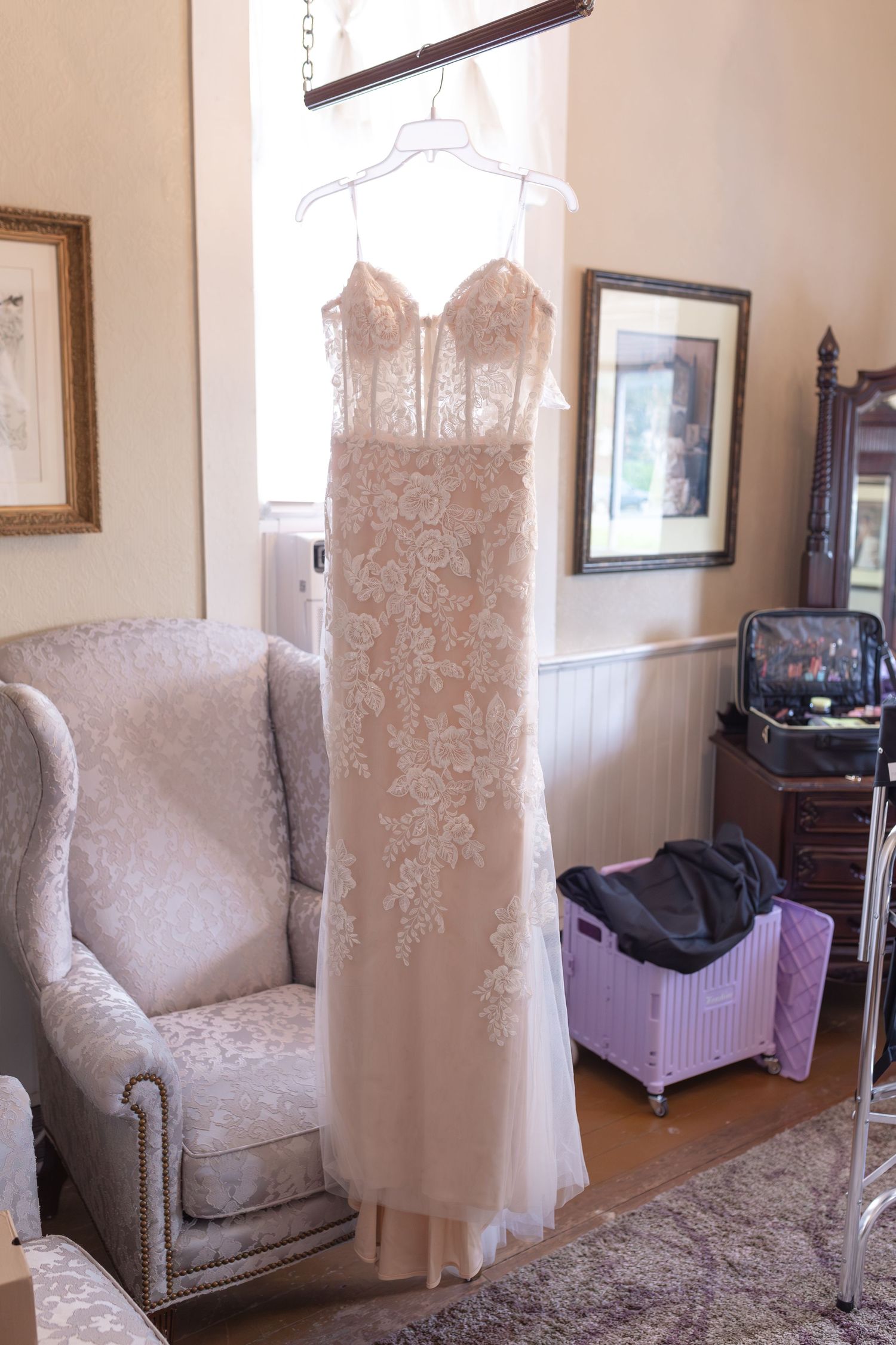 Elegant lace wedding dress hanging on a hanger near a window in a vintage-styled room.